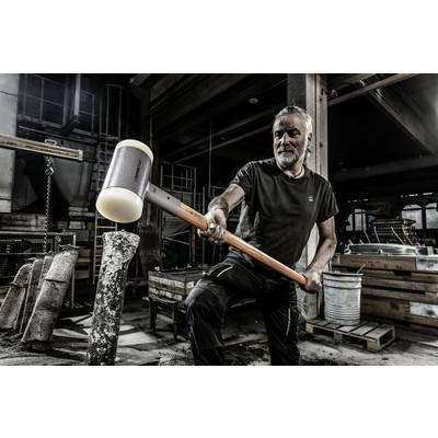 Ein Mann schlägt mit einem großen Hammer auf einen Steinblock in einer industriellen Werkstattumgebung. Er trägt schwarze Arbeitskleidung.