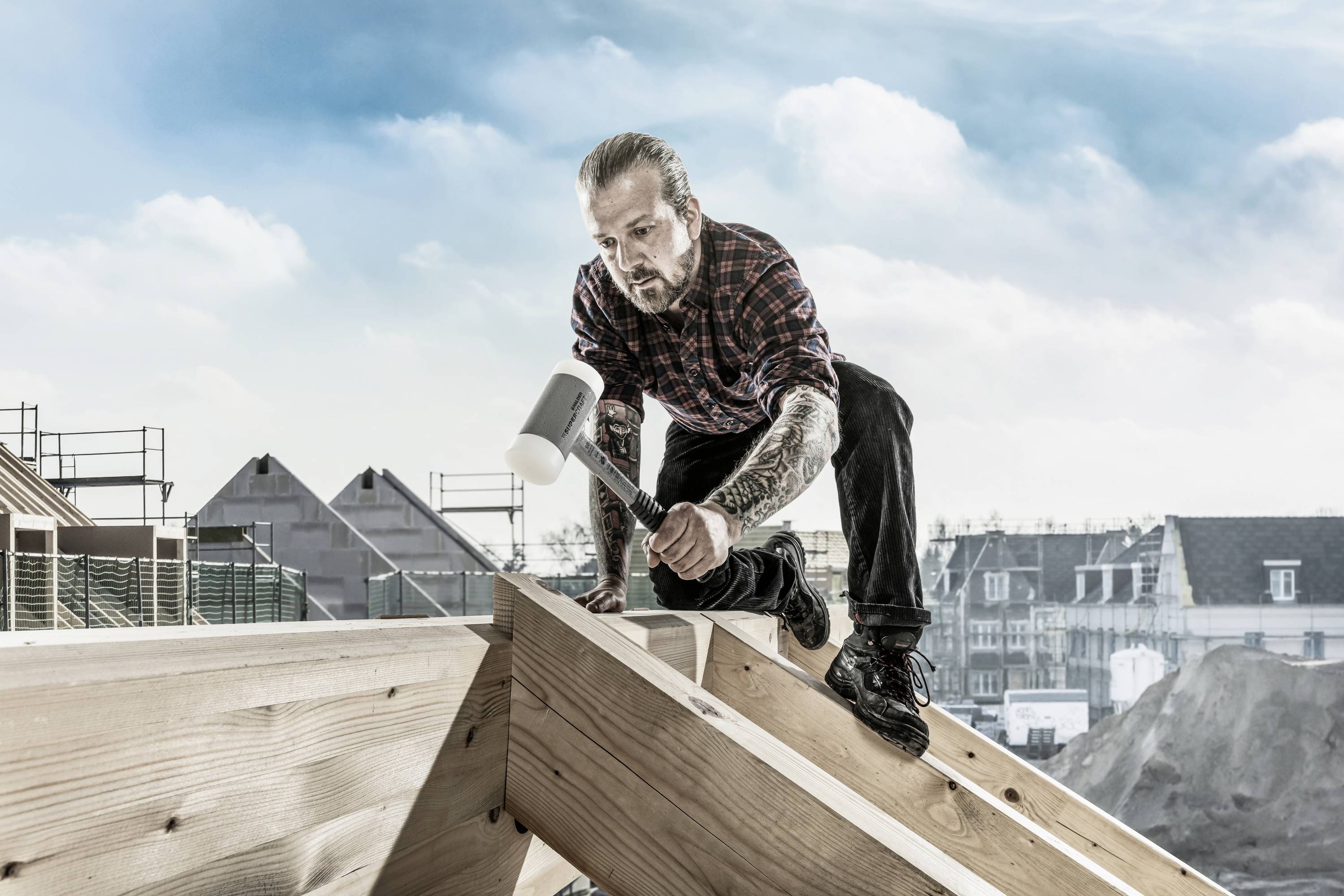 Ein Handwerker mit tätowierten Armen kniet auf einem Holzdach und benutzt einen Hammer, um die Konstruktion zu bearbeiten.