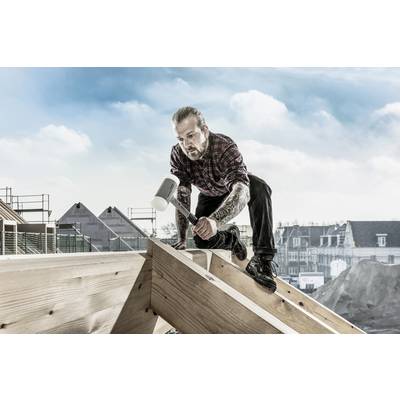 Ein Handwerker mit tätowierten Armen kniet auf einem Holzdach und benutzt einen Hammer, um die Konstruktion zu bearbeiten.