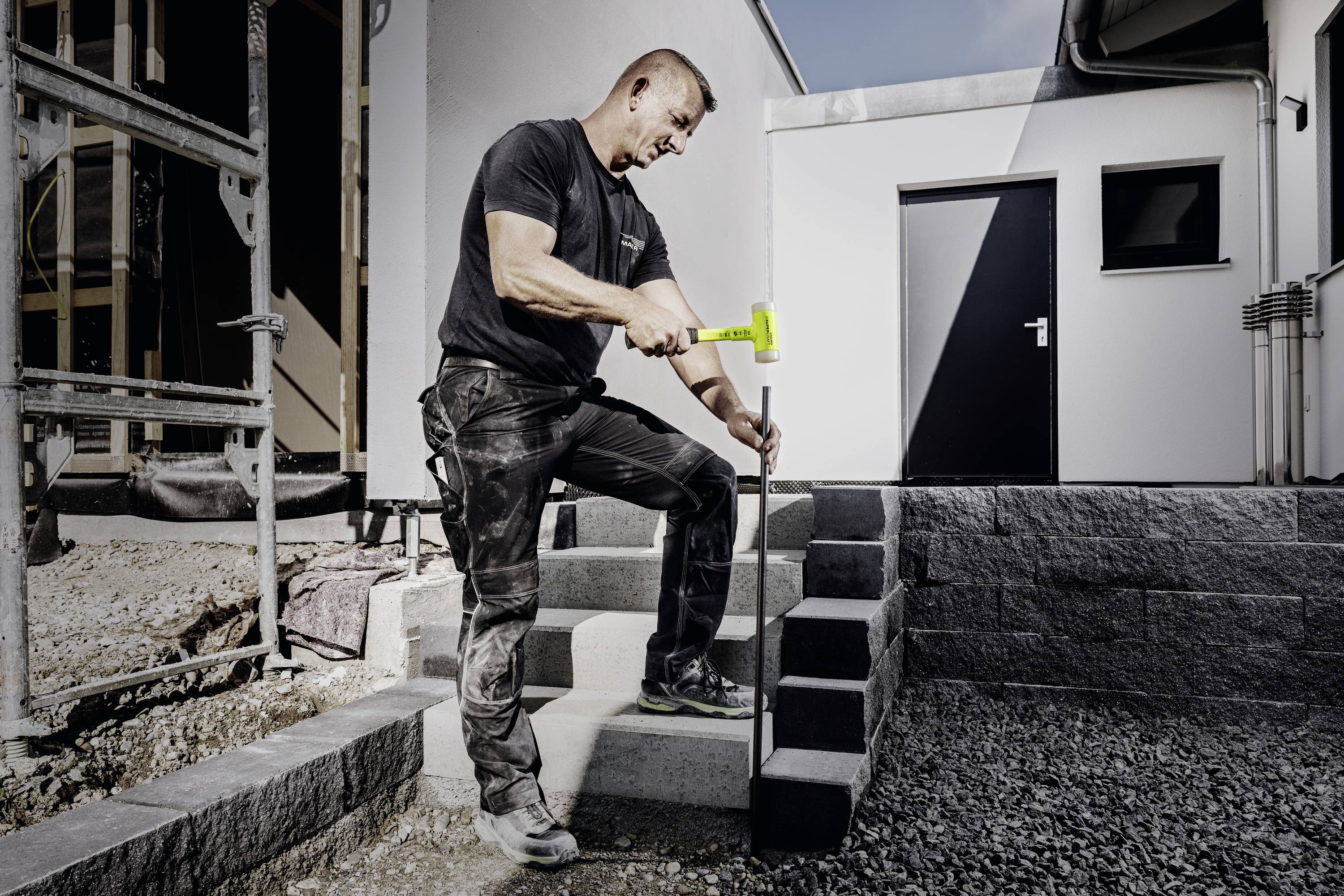 Ein Mann in Arbeitskleidung befestigt mit einem Hammer eine Steinverkleidung an einer Treppe vor einem modernen Gebäude.