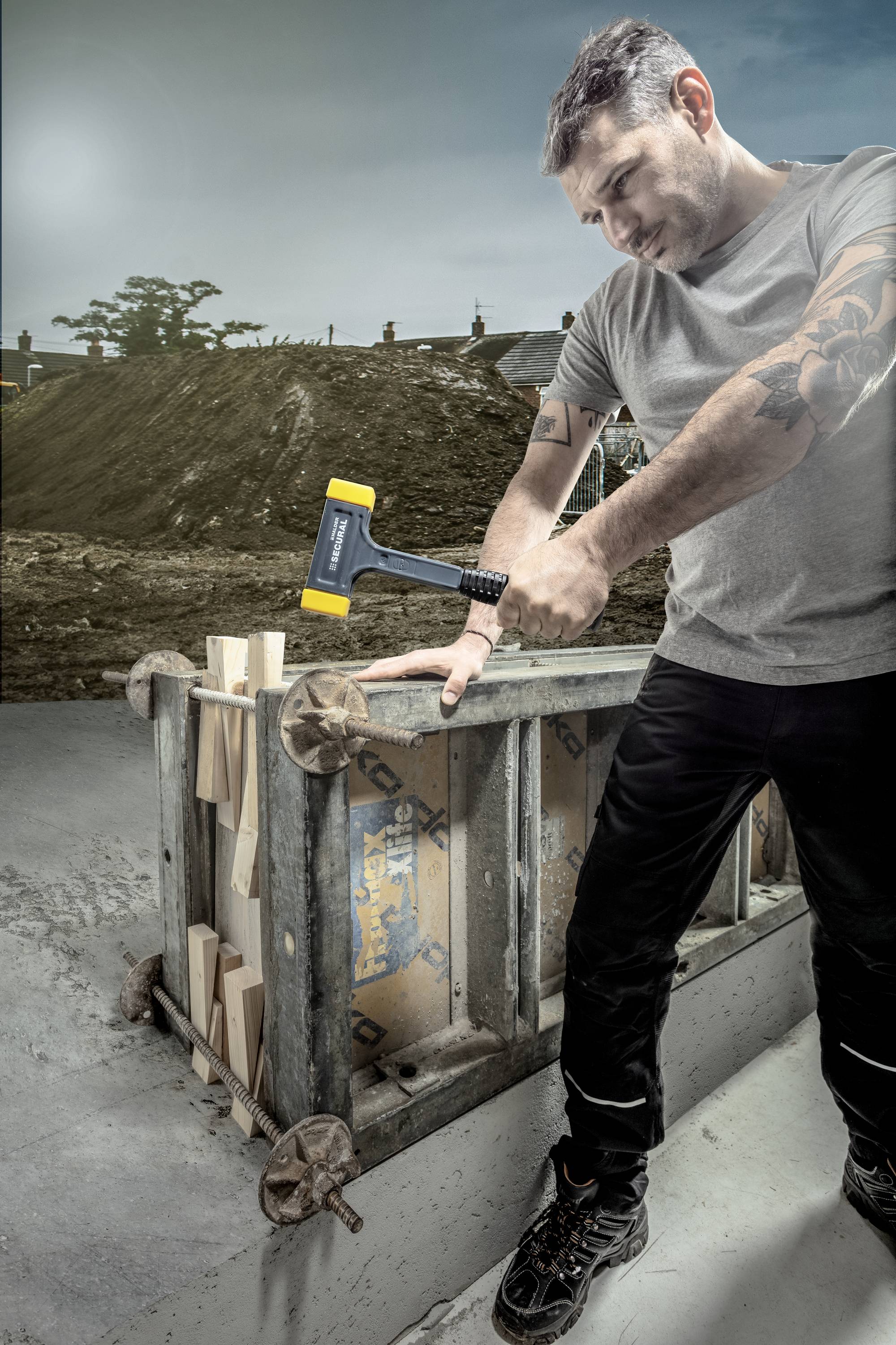 Ein Mann mit Tätowierungen benutzt einen Hammer, um Holzstücke in eine Metallform zu schlagen. Im Hintergrund ist eine Baustelle zu sehen.