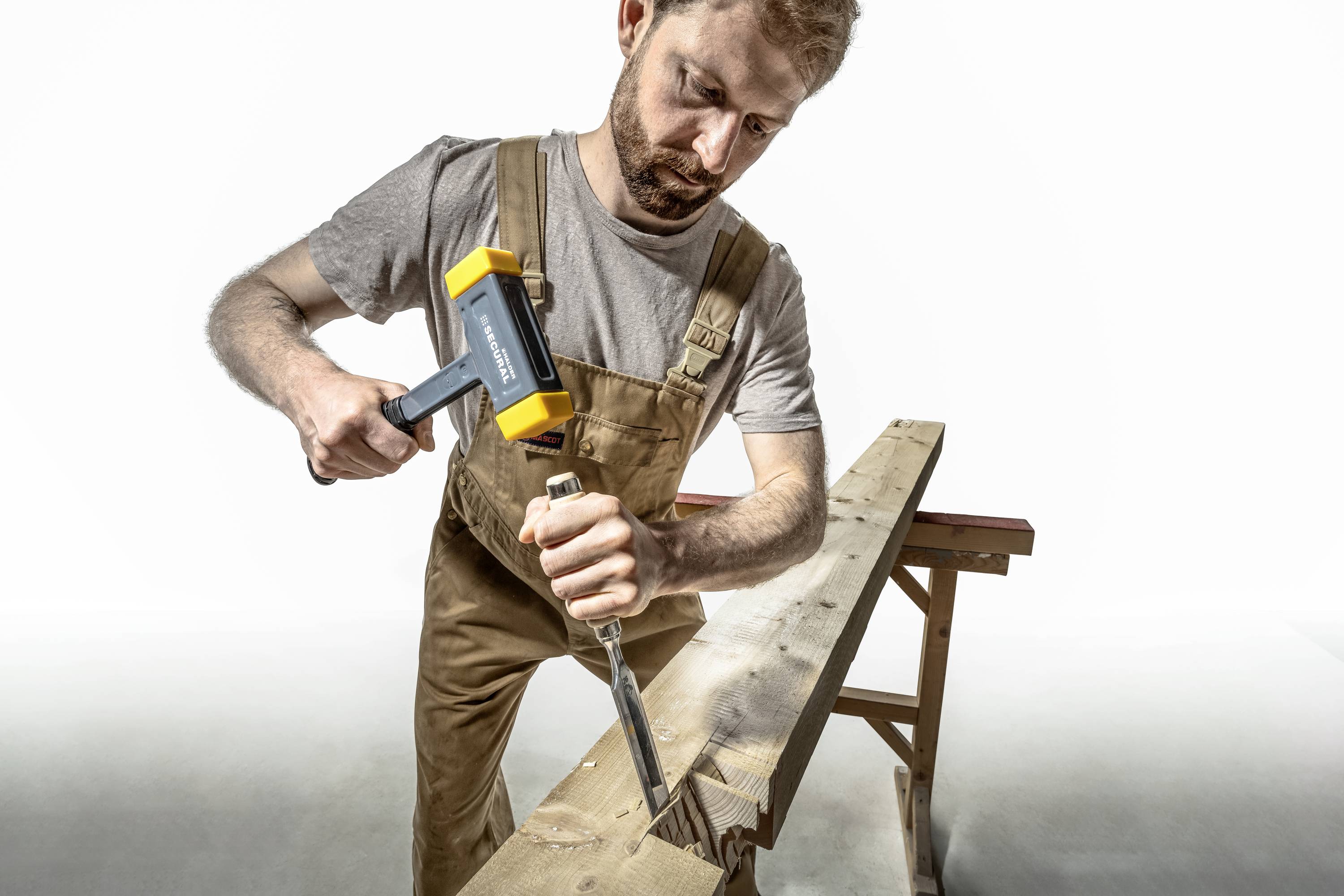 Ein Mann, der in Arbeitskleidung mit einem Hammer und Meißel an einem Holzstück arbeitet.