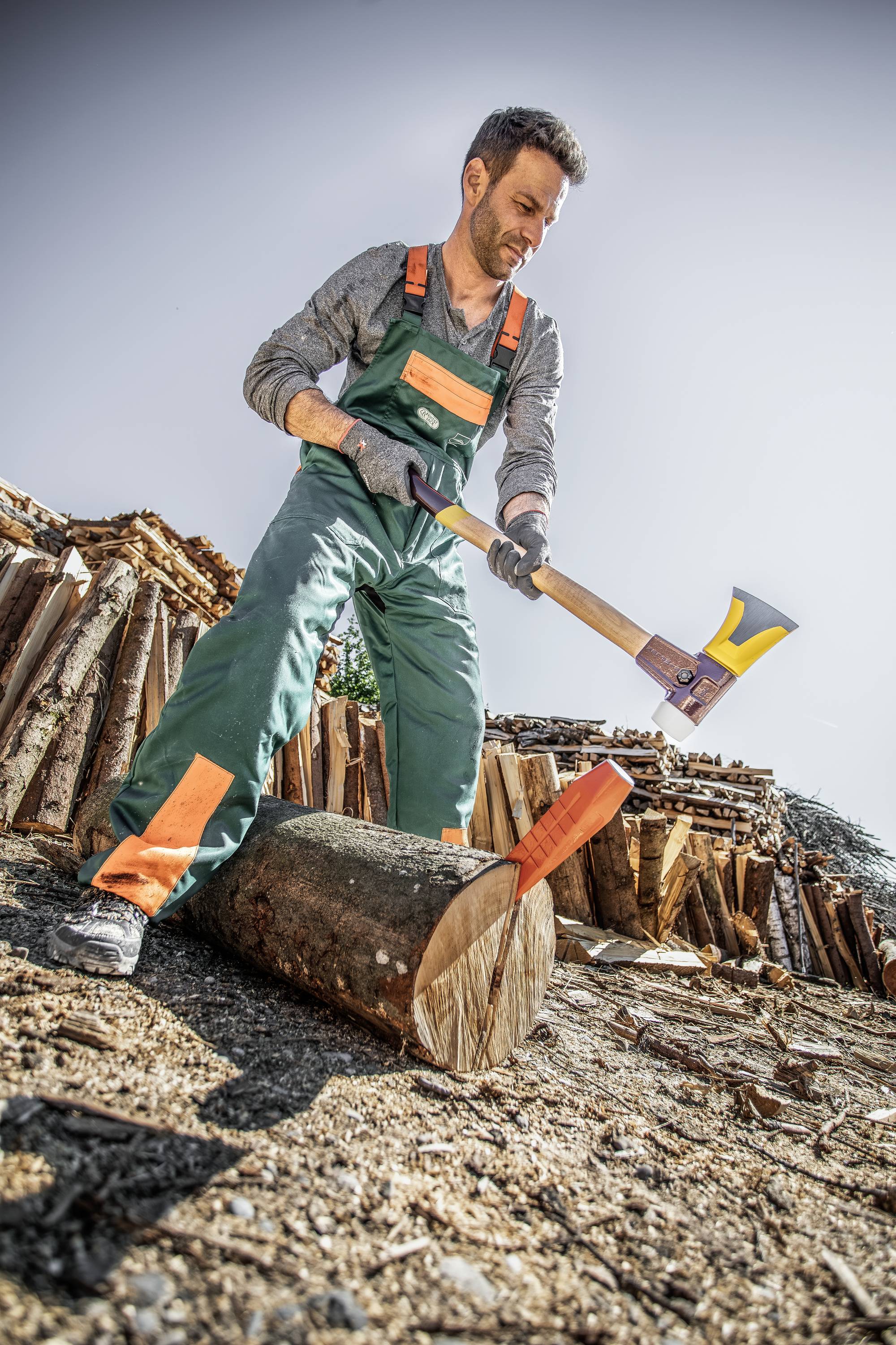 Ein Mann in Arbeitskleidung spaltet Holz mit einer Axt im Freien. Im Hintergrund sind gestapelte Holzscheite zu sehen.