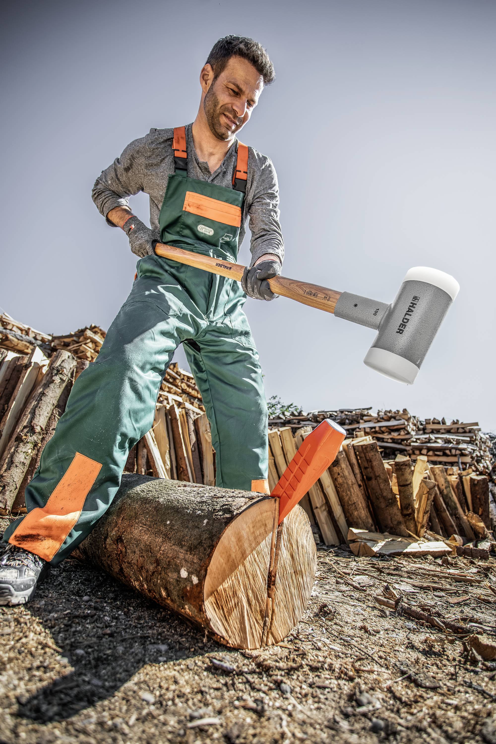 Ein Mann in grüner Arbeitskleidung spaltet mit einem großen Hammer einen Holzscheit, im Hintergrund aufgestapelte Holzstämme.