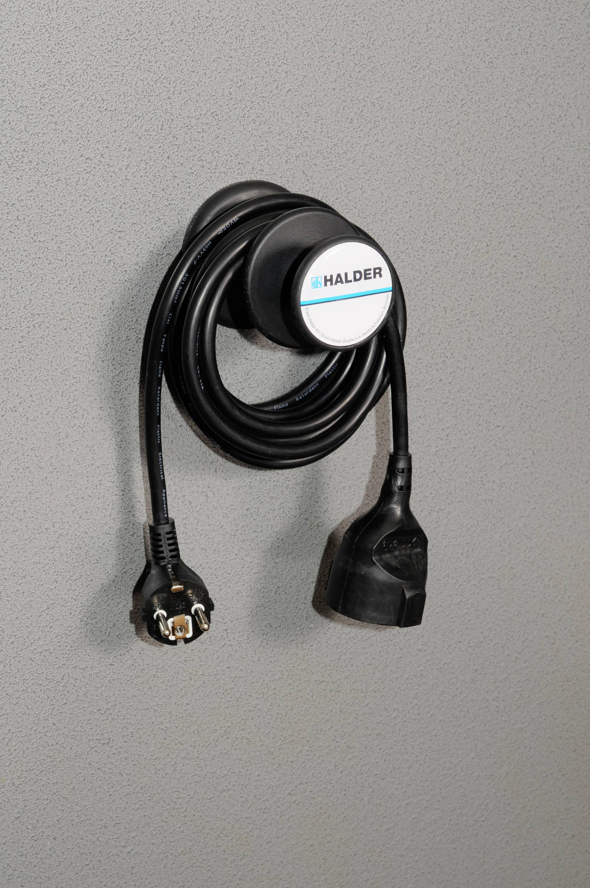 Ein Kabel mit rundem Steckerhalter an einer grauen Wand angebracht. Der Halter hat einen Aufkleber mit der Aufschrift 'HALDER'.