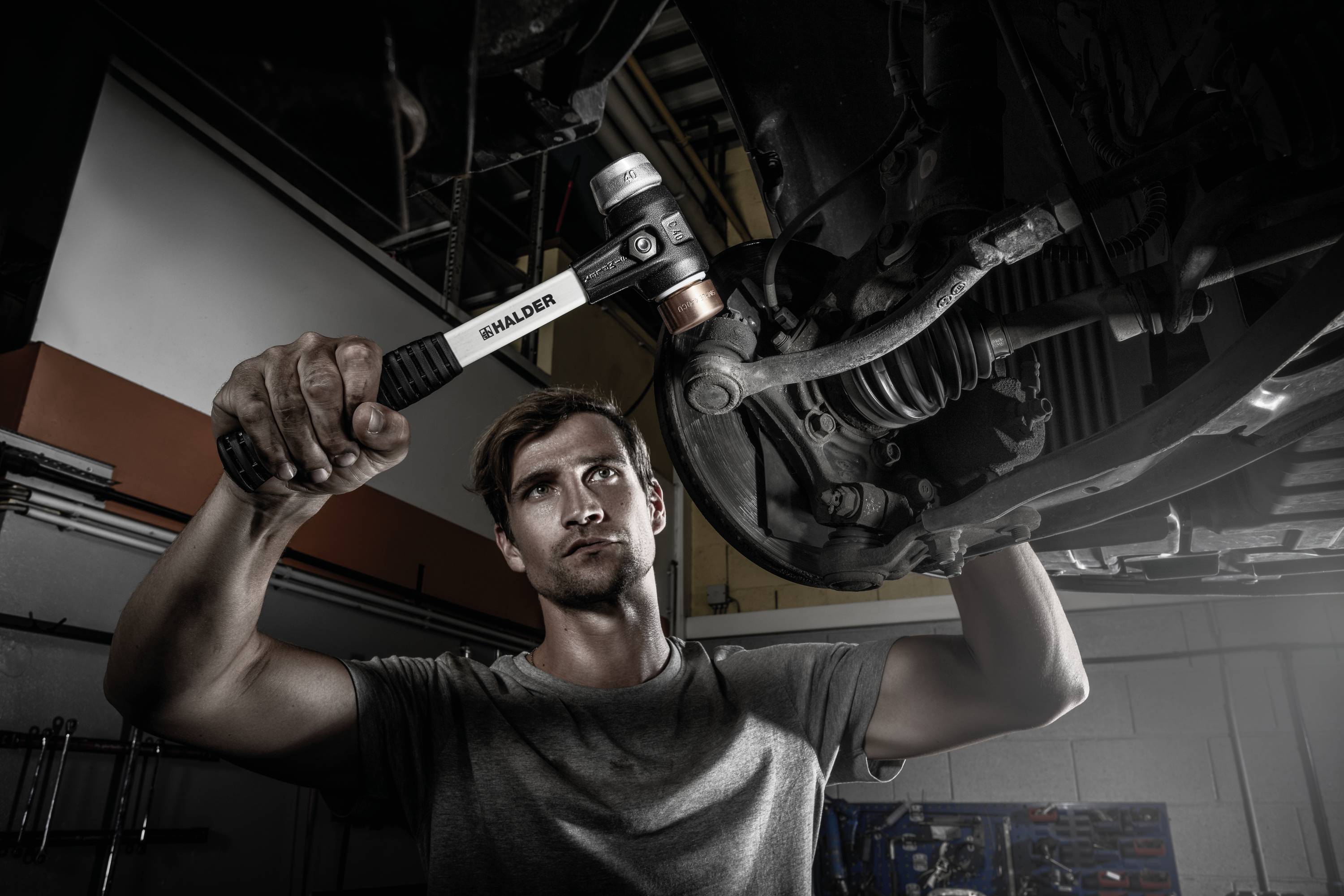 Mechaniker in grauem T-Shirt repariert Auto unter der Hebebühne, fokusiert auf Arbeit. Schraubenschlüssel in der Hand, Arbeitsumgebung sichtbar.