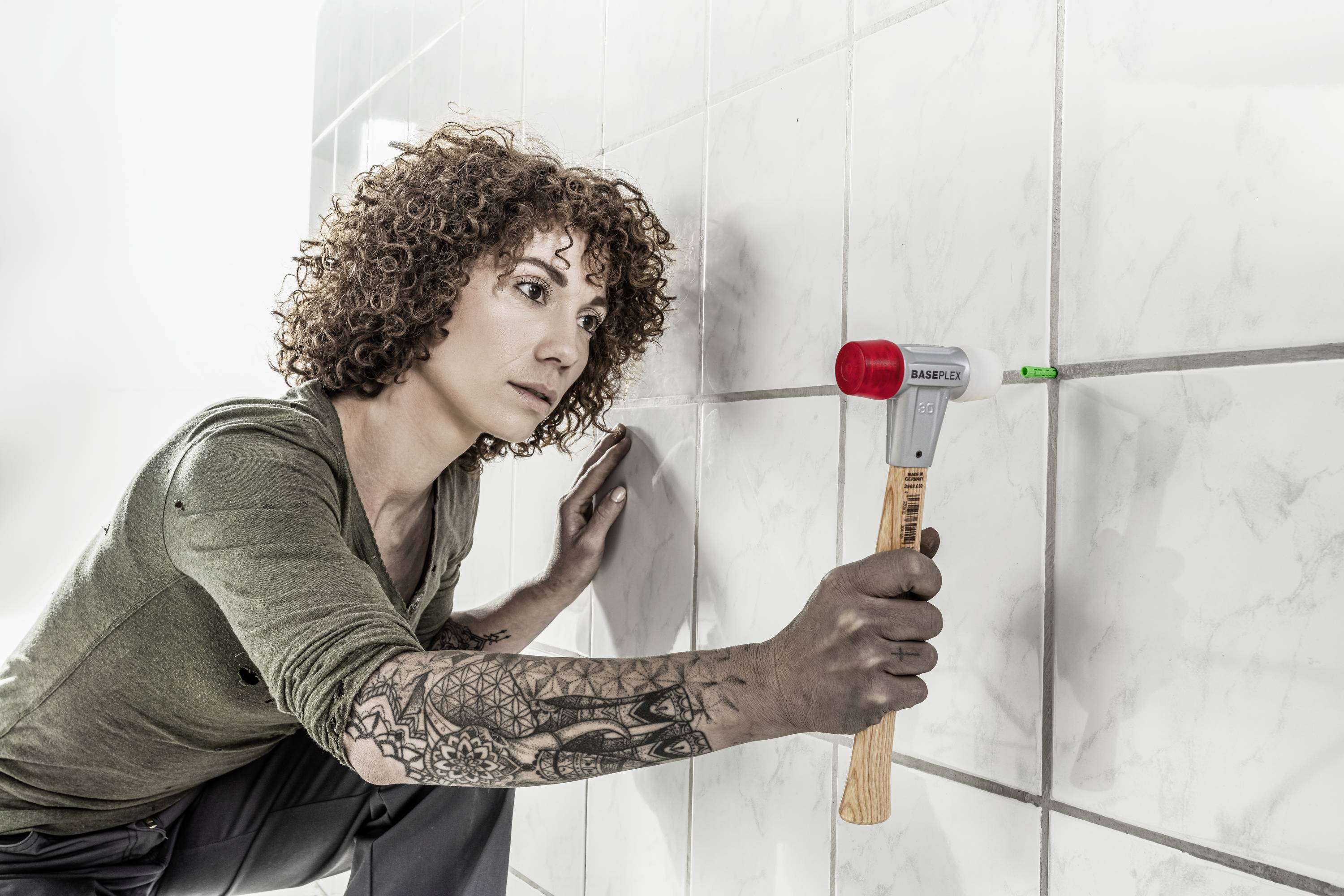 Person mit lockigem Haar benutzt einen Hammer, um einen grünen Stift in eine Wand mit weißen Fliesen zu schlagen. Tätowierter Arm sichtbar.