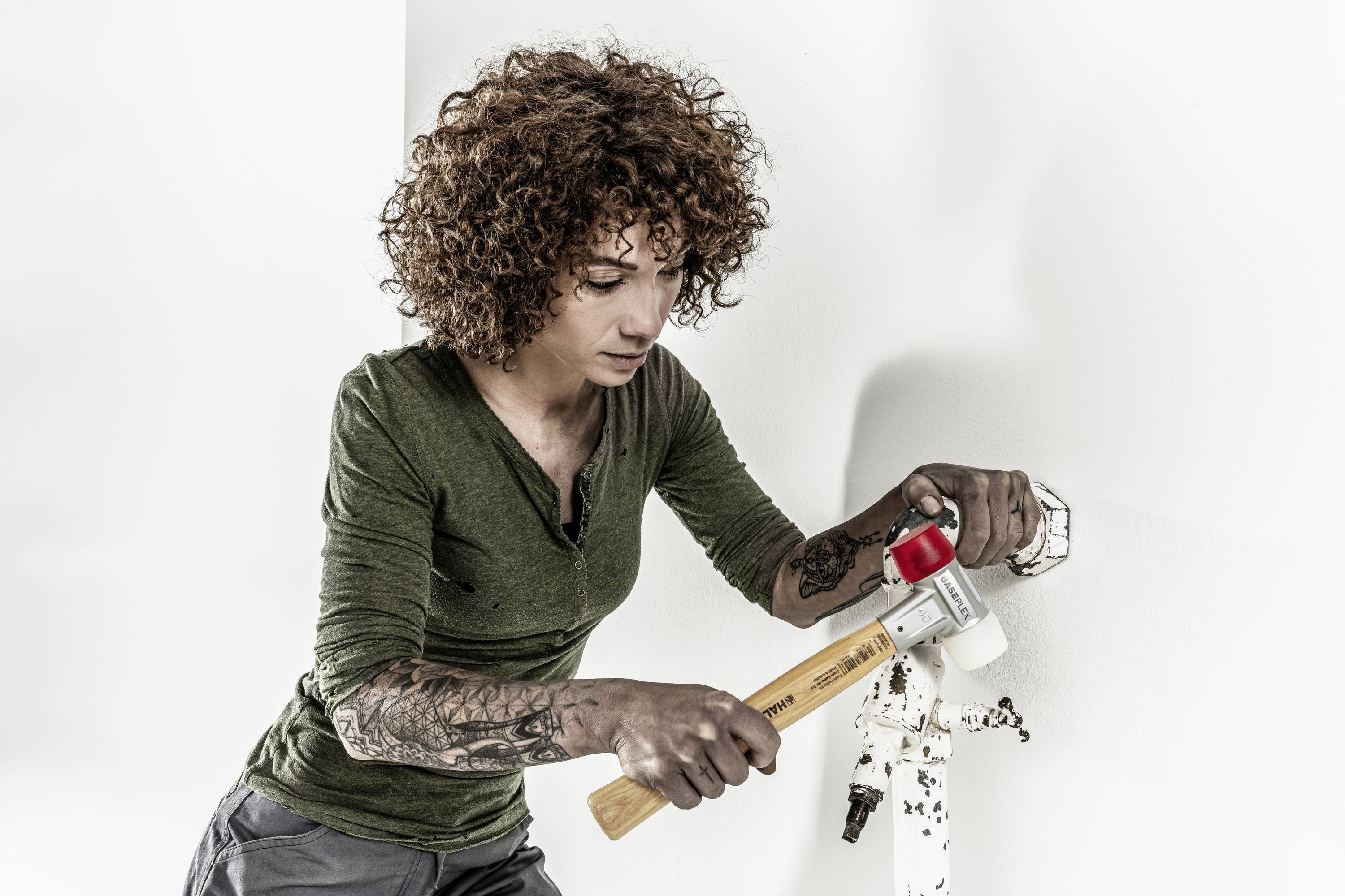 Eine Person mit lockigem Haar benutzt einen Hammer, um ein Rohr zu reparieren. Es fließt ein wenig Wasser aus dem Rohr.