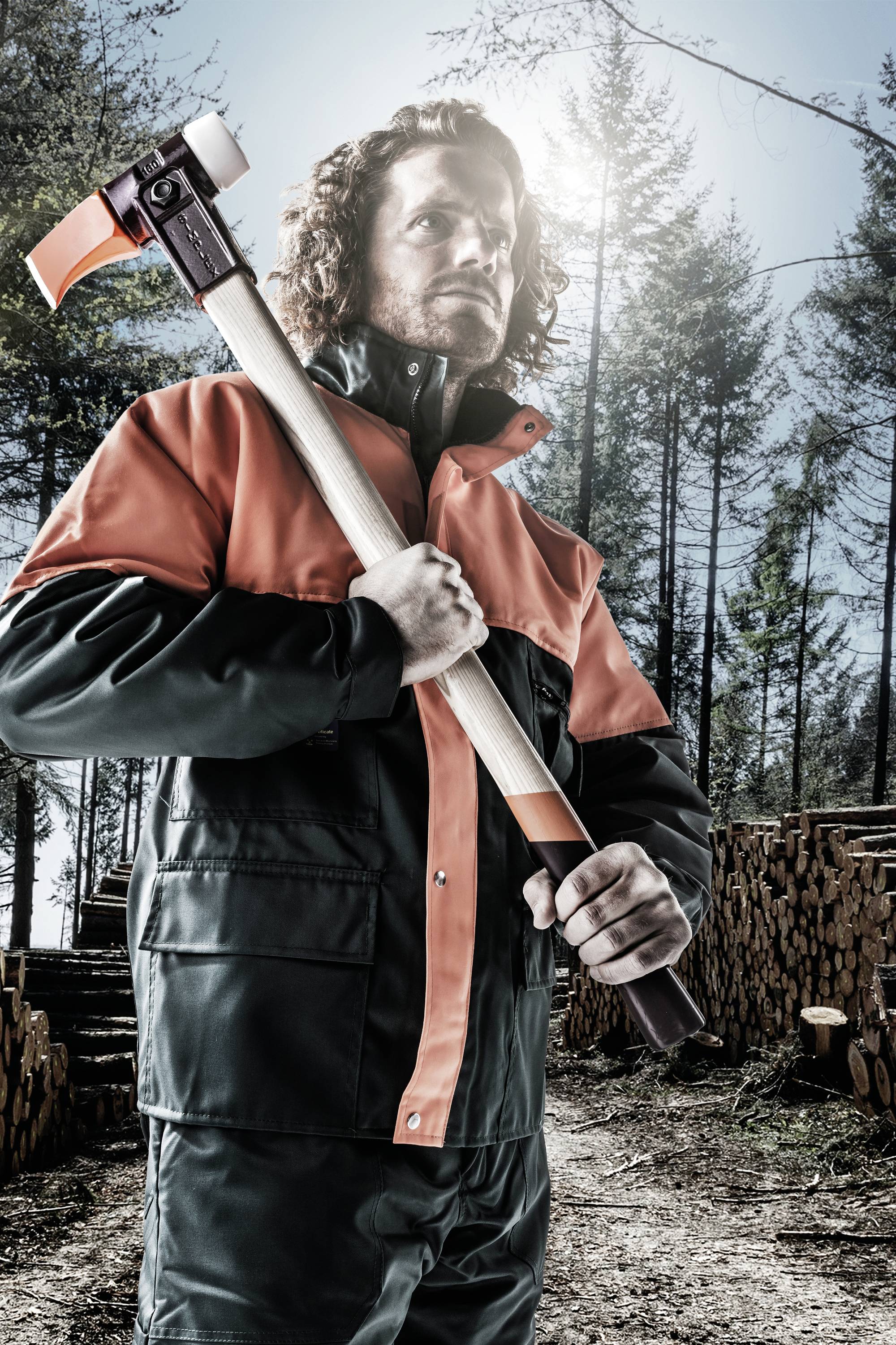 Arbeiter im orange-schwarzen Anzug steht im Wald und hält eine Axt, im Hintergrund gestapelte Holzscheite und Bäume.