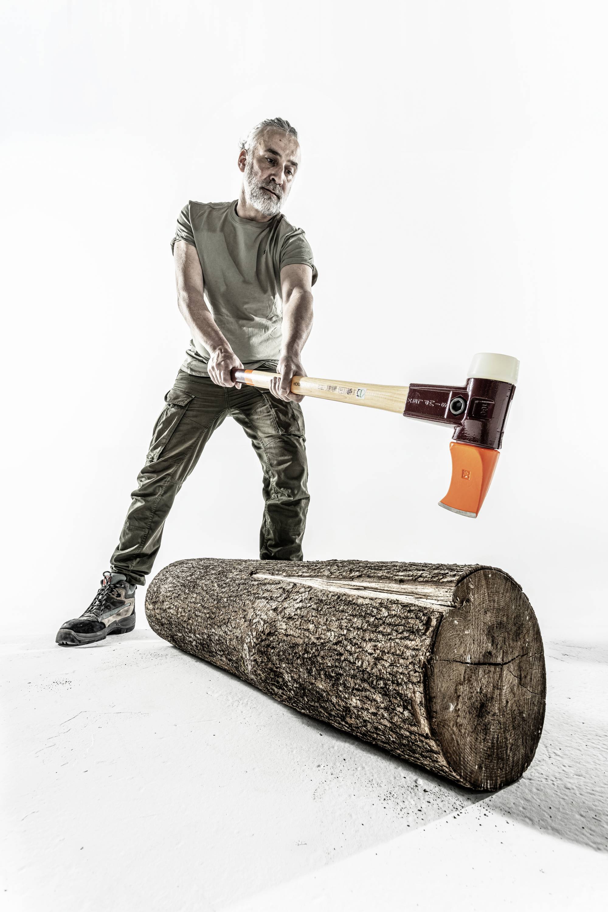 Ein Mann mit Bart spaltet mit einer Axt einen Holzstamm. Er trägt ein khakifarbenes T-Shirt und Hosen, steht auf einem weißen Hintergrund.