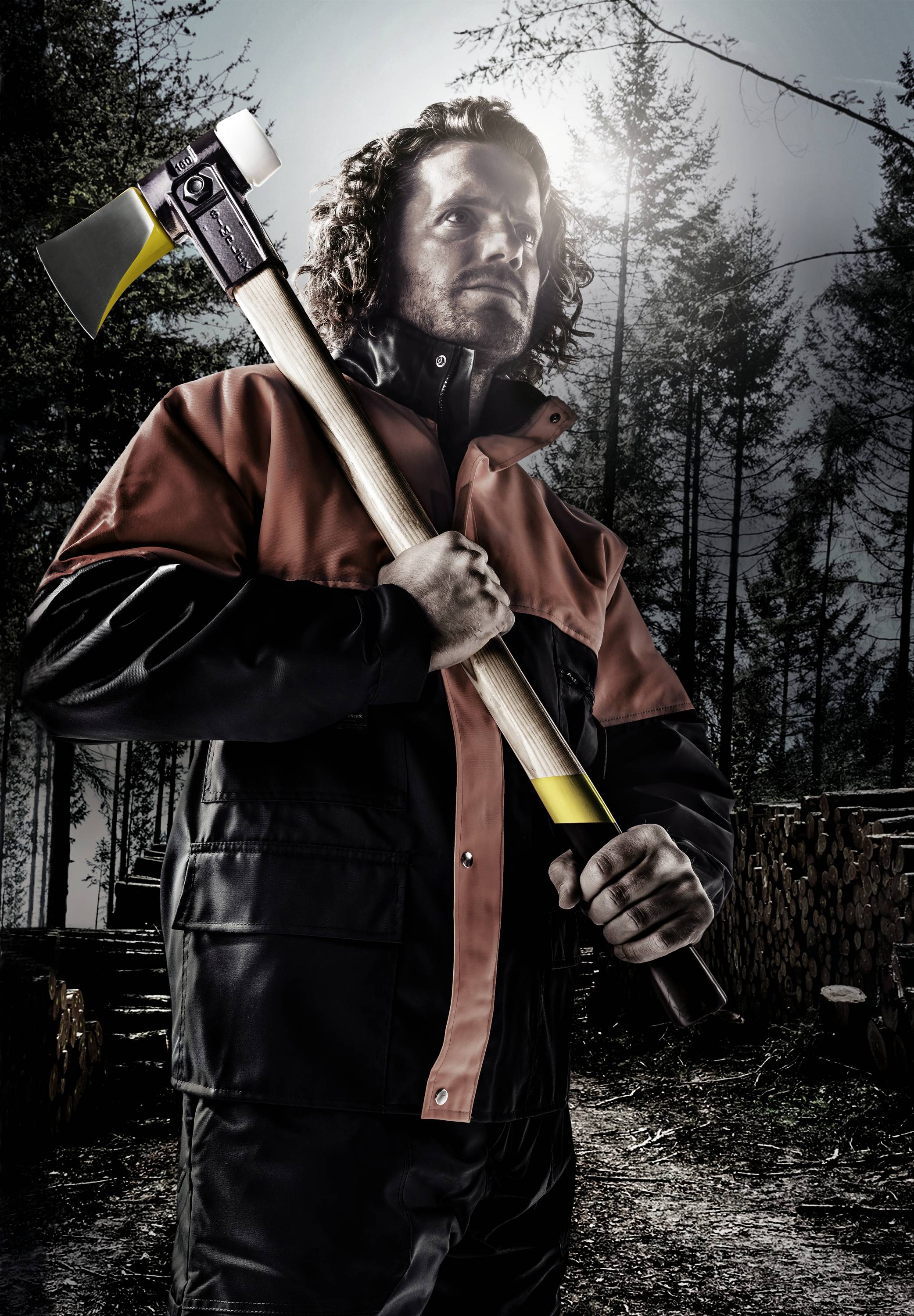 Ein Mann in Schutzkleidung hält eine Axt in einem Wald. Umgeben von Bäumen und Holzstapeln, wirkt er bereit für Forstarbeiten.