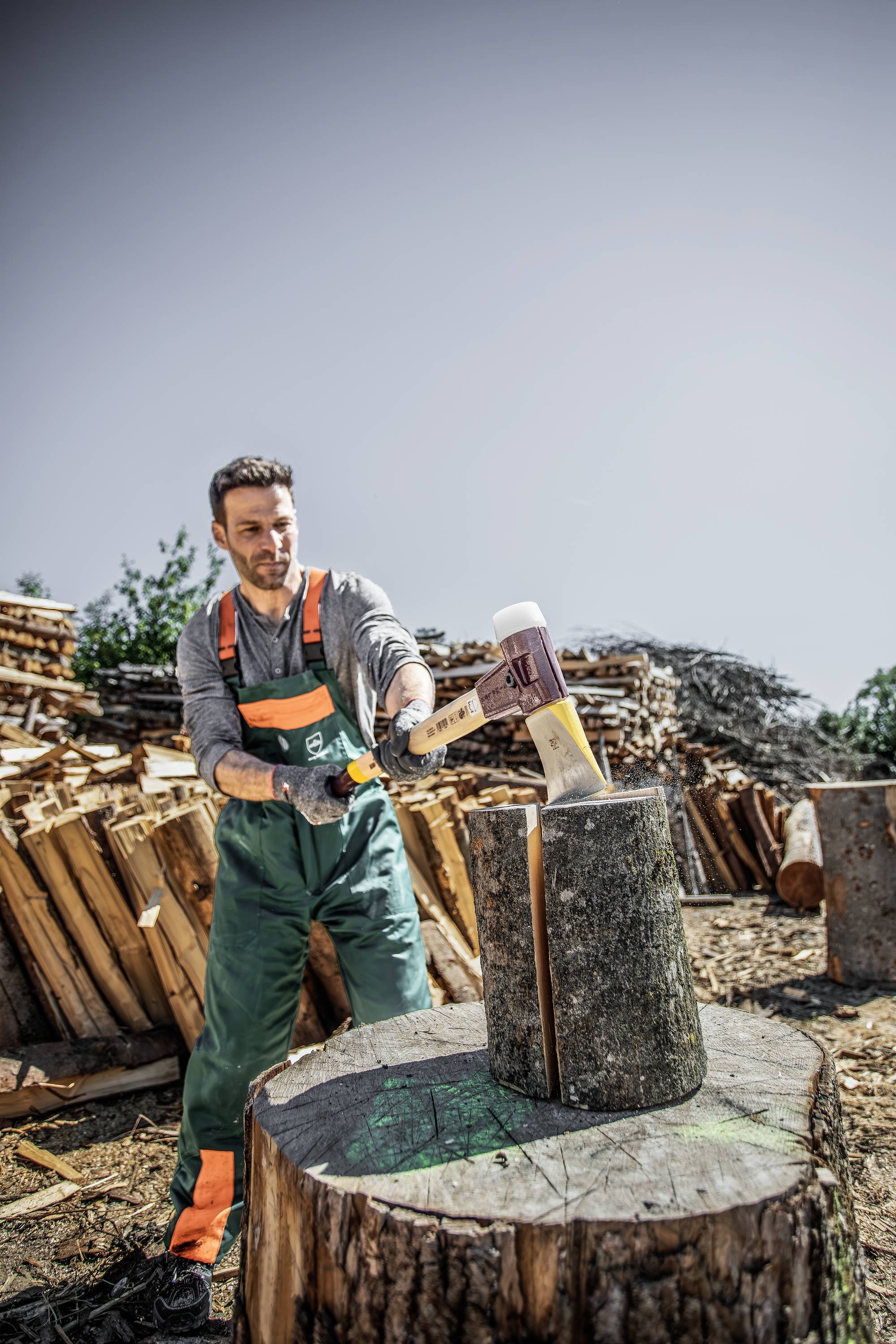 Ein Mann in Arbeitskleidung spaltet mit einer Axt Holzscheite im Freien. Im Hintergrund sind gestapelte Holzstapel zu sehen.