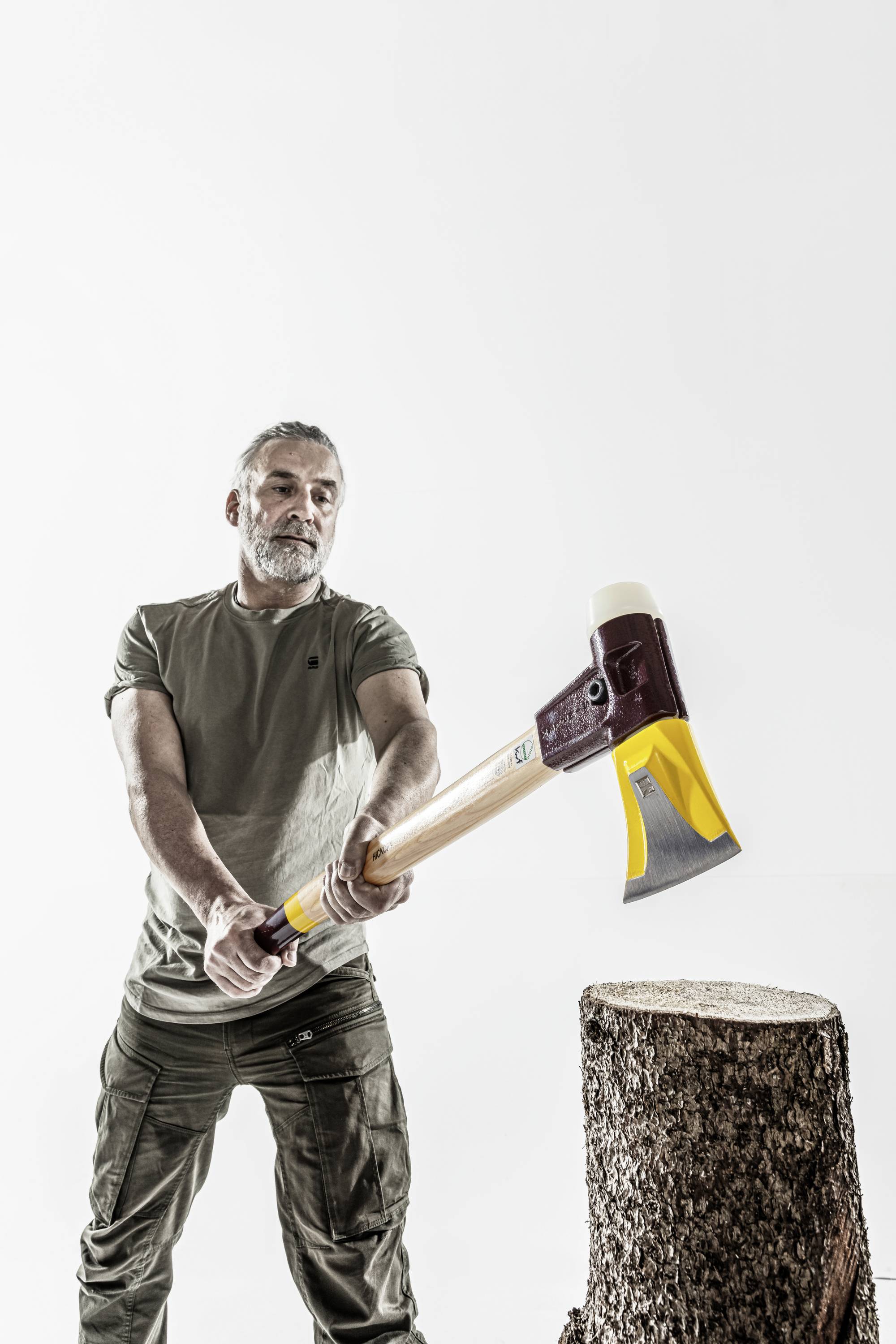 Ein Mann schwingt eine Axt, um einen Baumstumpf zu spalten. Er trägt ein T-Shirt und steht vor einem neutralen Hintergrund.