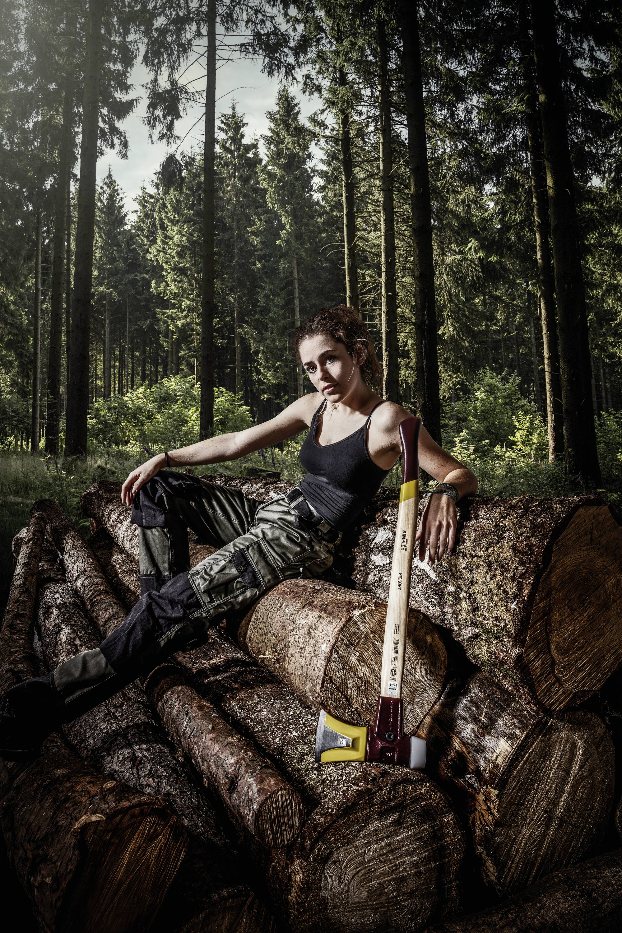 Eine Person sitzt lässig auf einem Stapel Baumstämme in einem Wald. Eine Axt lehnt an den Stämmen. Dichte Bäume im Hintergrund.