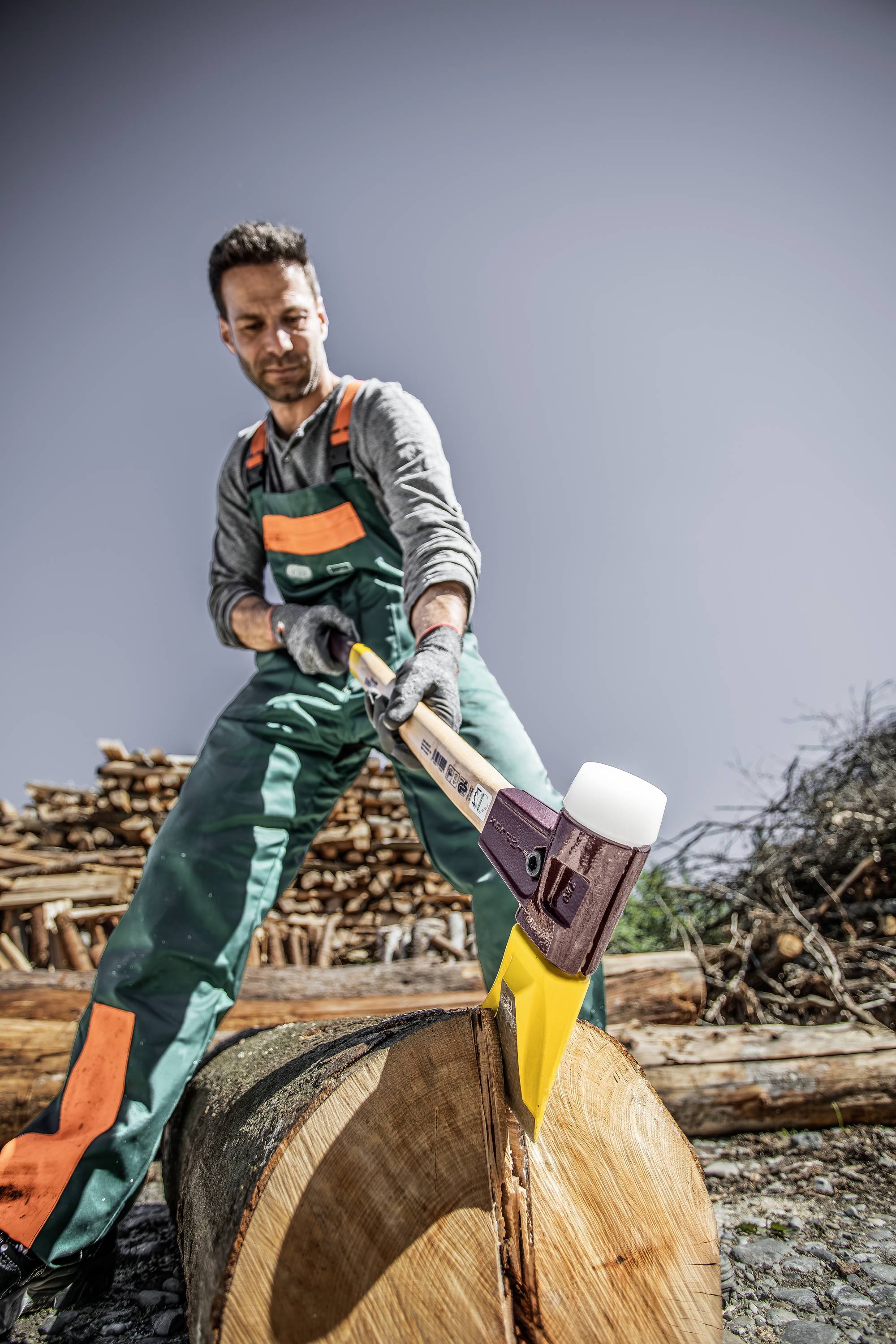 Ein Mann in Arbeitskleidung spaltet draußen mit einer Axt einen großen Holzblock. Im Hintergrund sieht man Holzstapel und Äste.