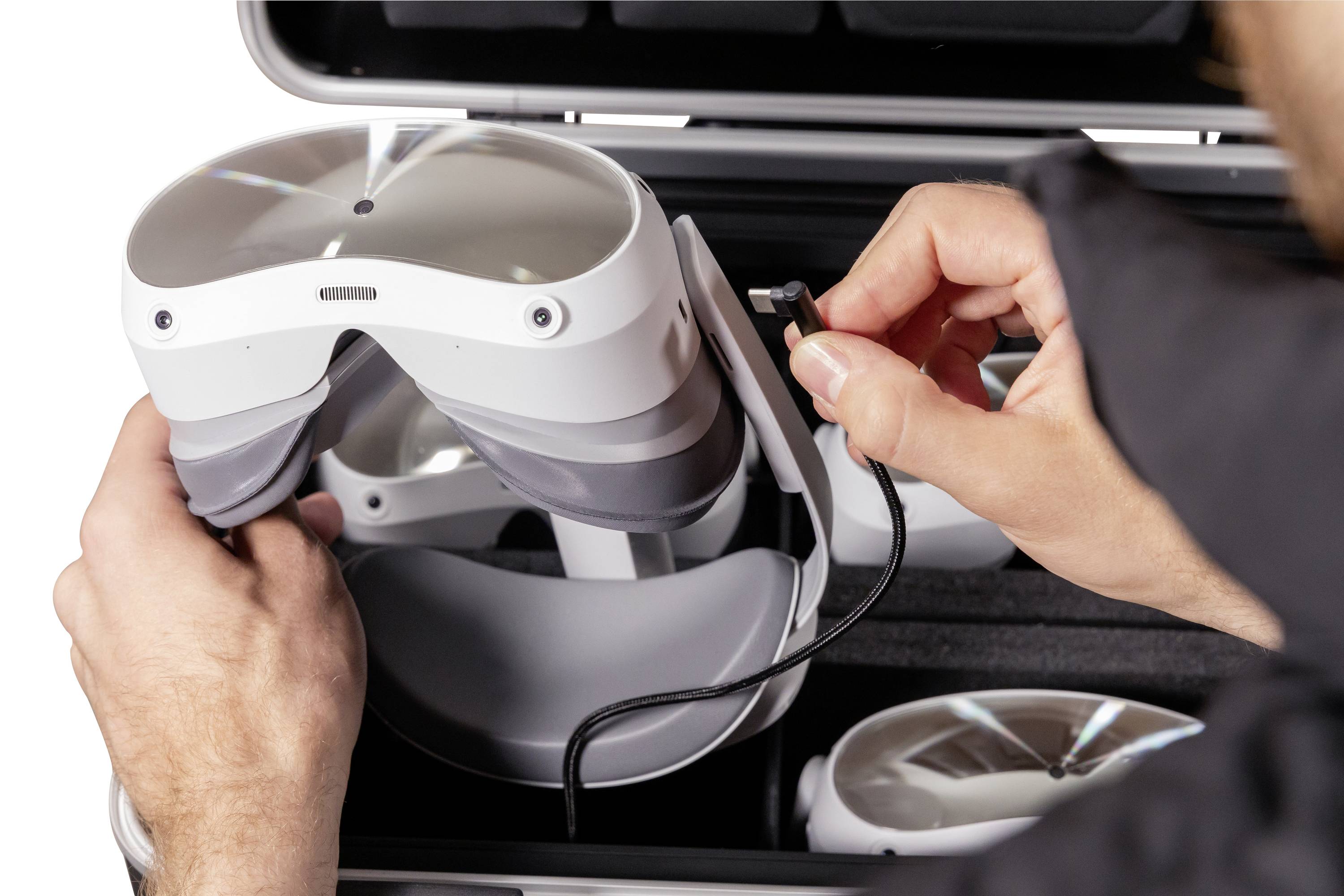 Eine Person verbindet ein VR-Headset mit einem Kabel, vermutlich zum Laden oder zur Datenübertragung. Das Headset liegt in einem offenen Koffer.