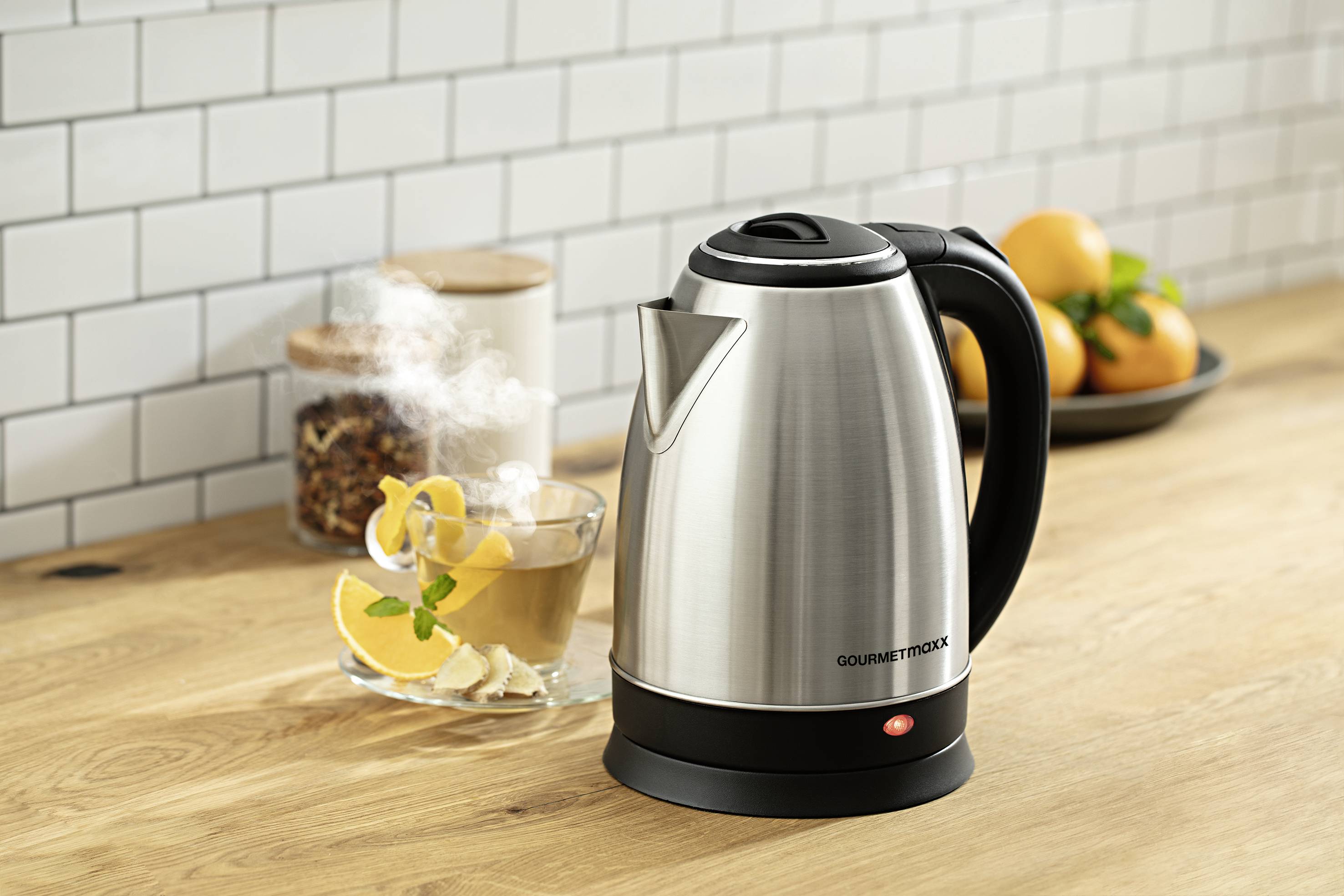 Ein silberner Wasserkocher auf einem Holztisch neben einer dampfenden Teetasse mit Zitrone und einem Teller mit Orangen im Hintergrund.