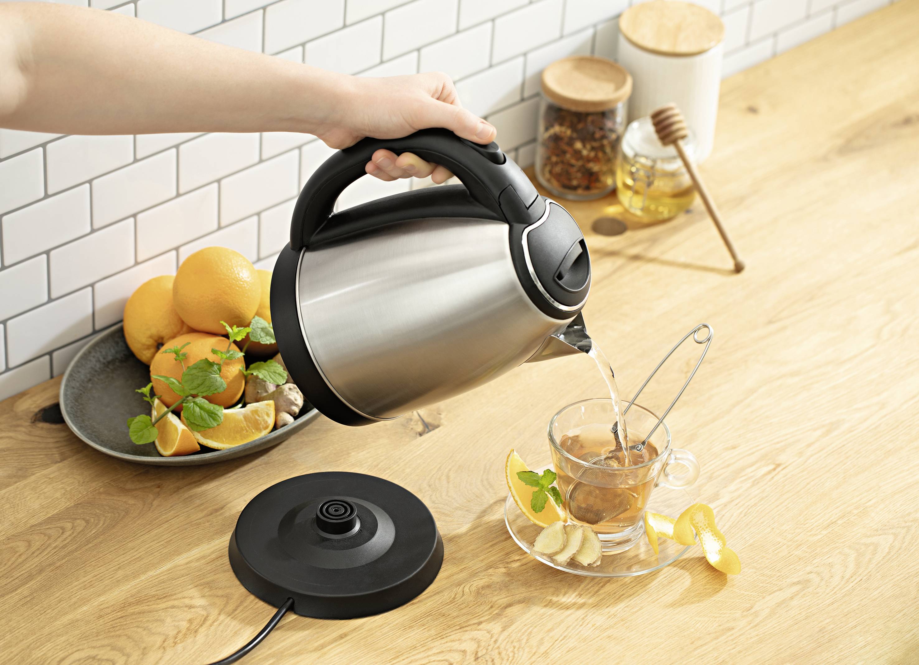 Eine Hand gießt heißes Wasser aus einem Wasserkocher in eine Teetasse. Im Hintergrund Obst, Gewürze und Dekoration auf einem Holztisch.