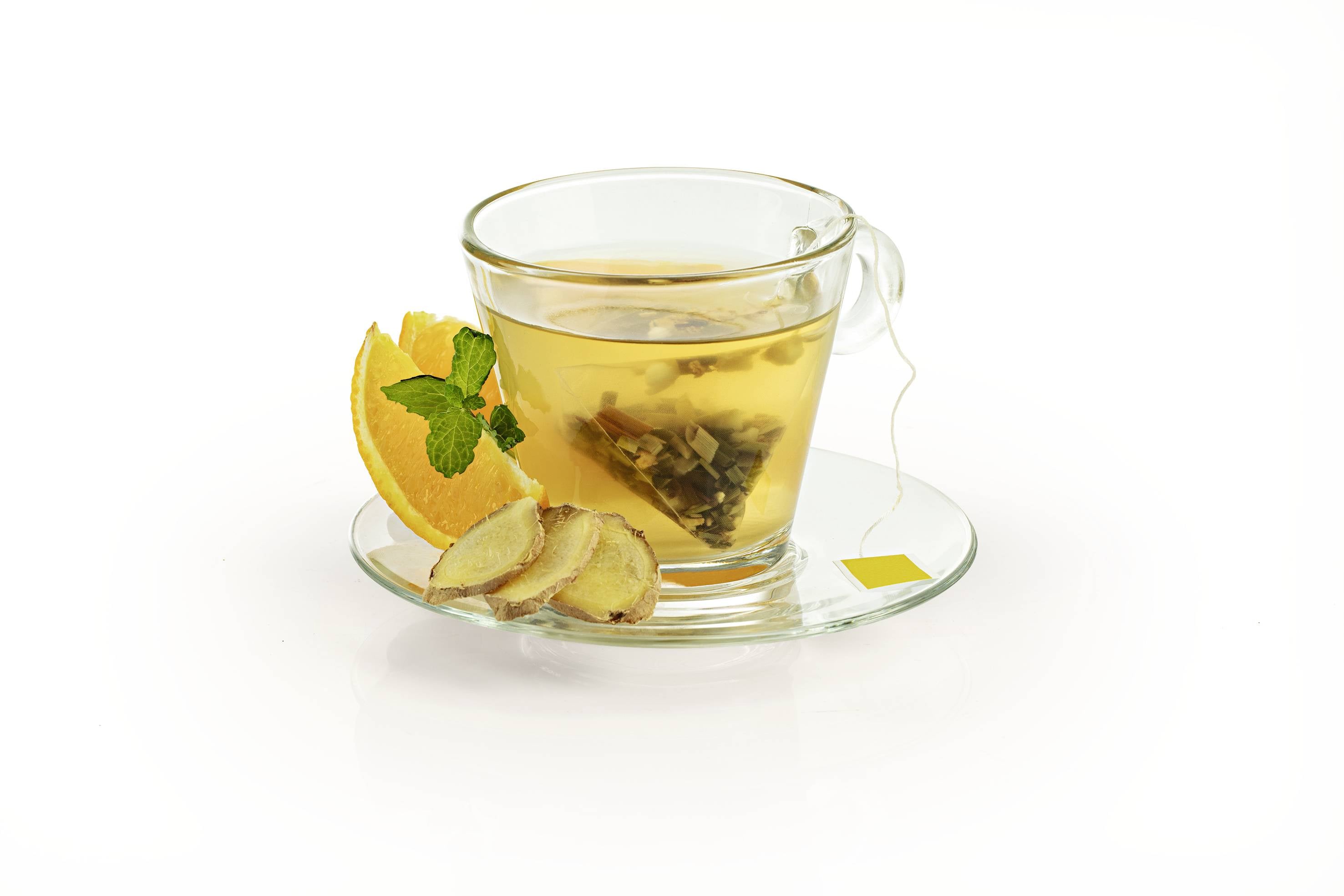 Ein Glas Tee mit einem Teebeutel, an einer Untertasse mit Orangenscheibe, Minzblatt und Ingwerscheiben, auf weißem Hintergrund.