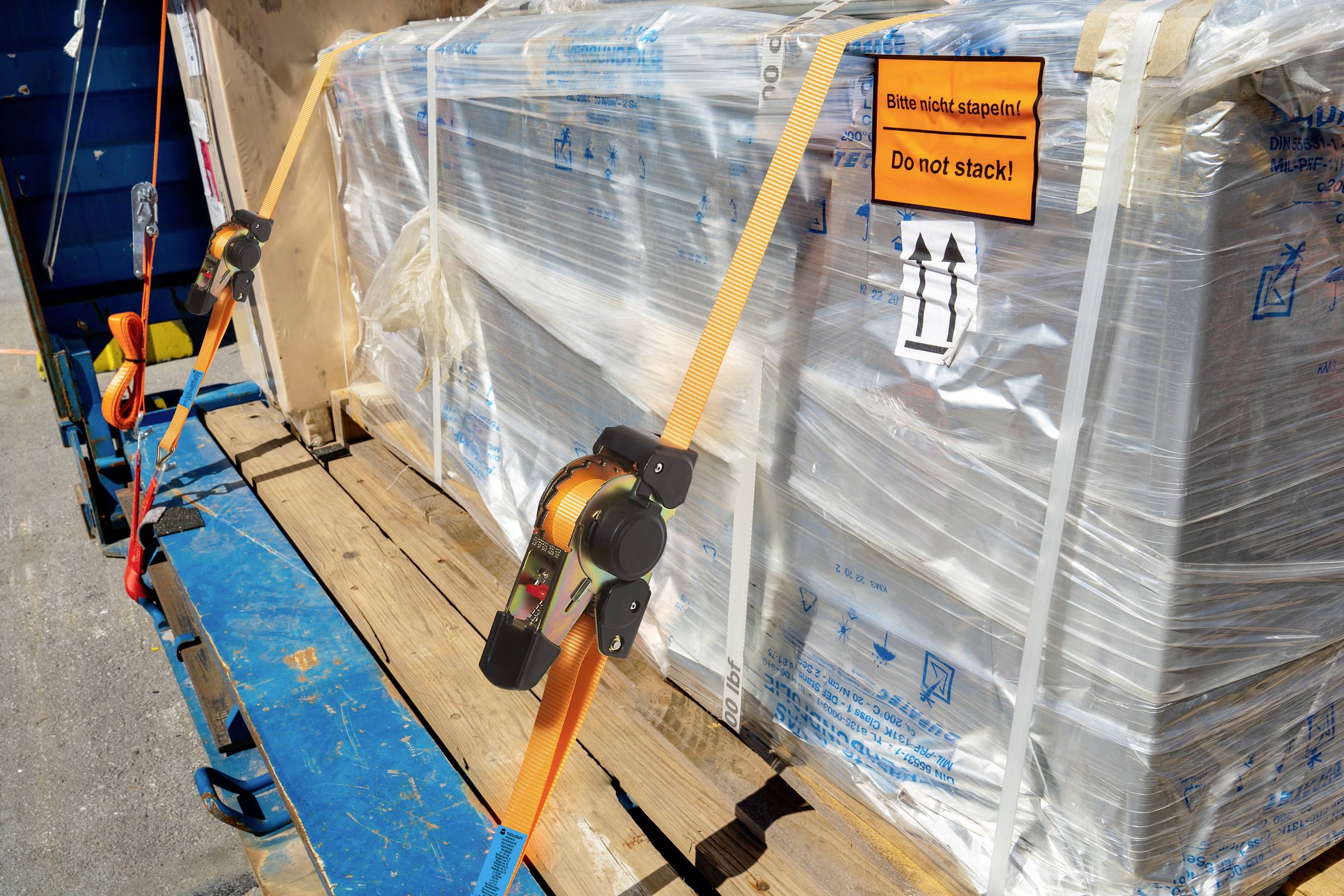 Eine in Folie verpackte Ladung ist mit orangefarbenen Gurten auf einem blauen Anhänger gesichert. Schild: 'Bitte nicht stapeln!'.