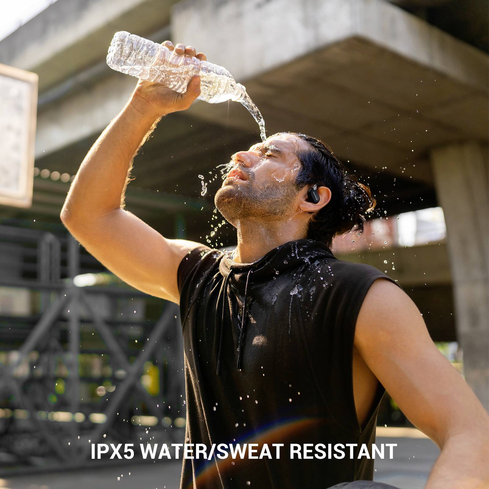 Ein Mann gießt Wasser aus einer Flasche über seinen Kopf. Text im Bild: 'IPX5 wasser-/schweißresistent'. Er steht unter einer Brücke.
