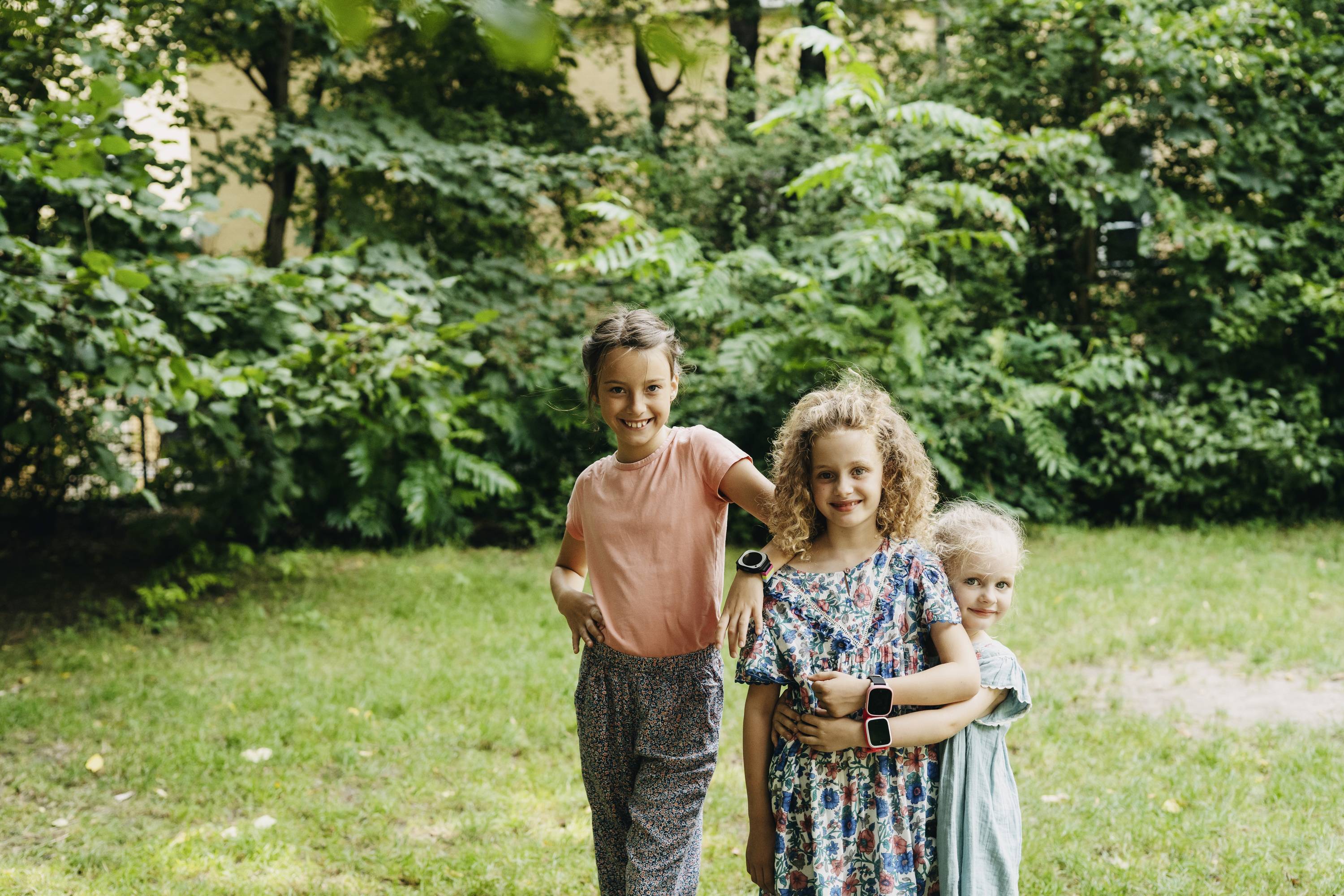 Drei Kinder stehen lächelnd im Garten; sie tragen bunte Kleidung und Armbänder. Im Hintergrund sind dichte grüne Büsche und Bäume.