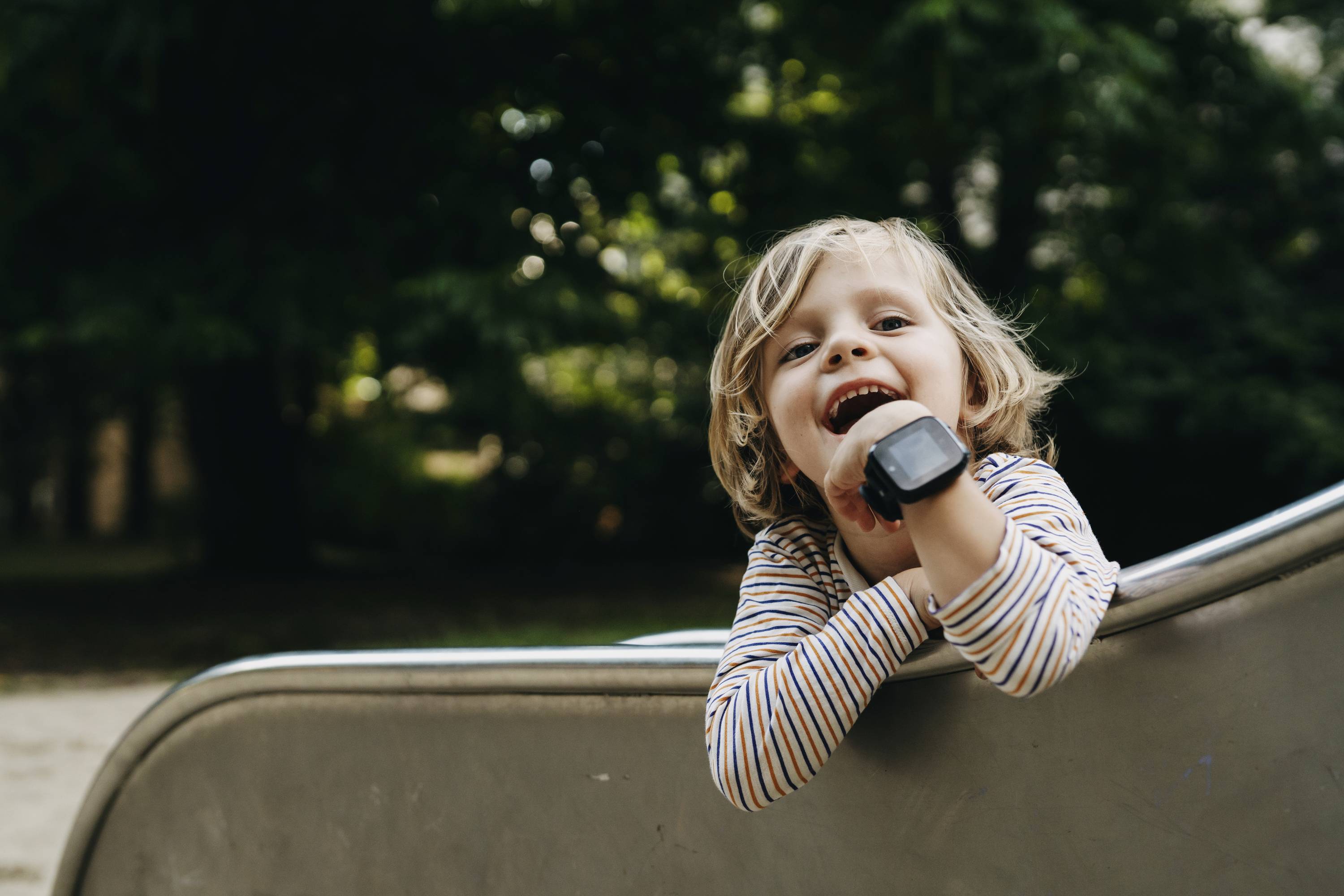 Ein lachendes Kind mit gestreiftem Oberteil lehnt sich über eine Rutsche auf einem Spielplatz. Pagettliche Stimmung im Hintergrund.