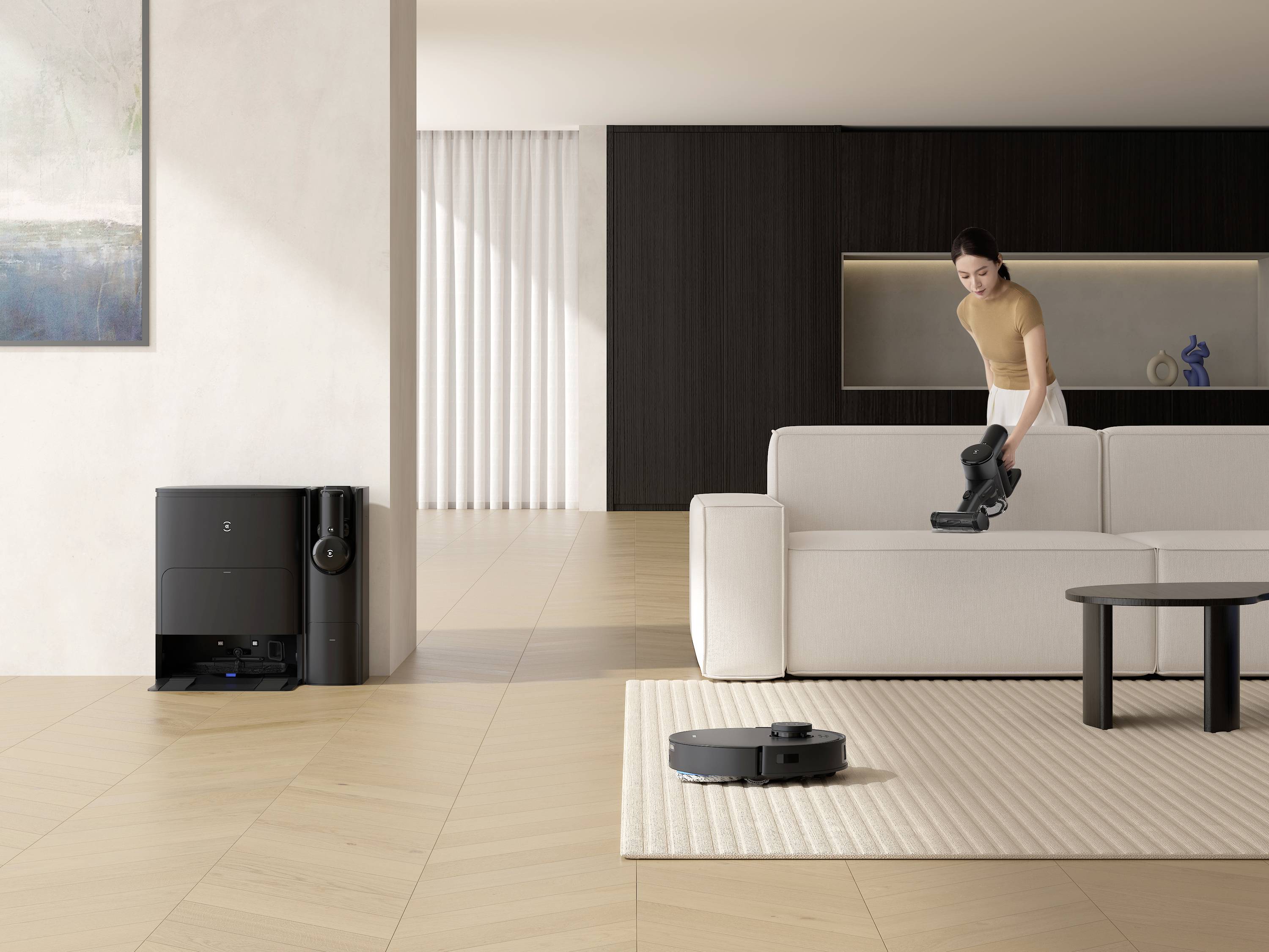 Eine Frau benutzt einen Handstaubsauger auf einem weißen Sofa in einem modernen Wohnzimmer, während ein Roboterstaubsauger den Boden reinigt.