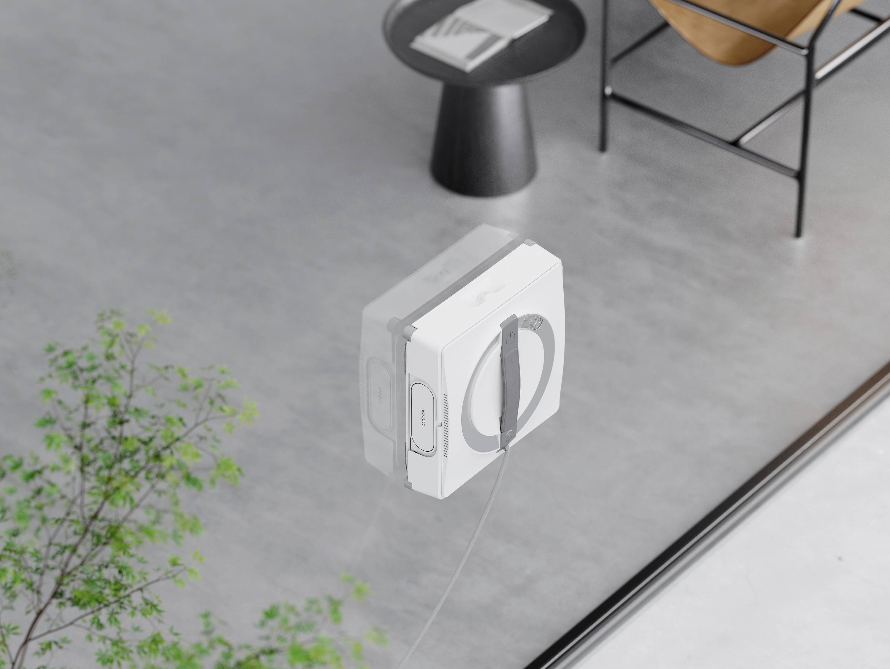 Ein quadratischer Fensterputzroboter reinigt eine große Glasscheibe in einem modernen, minimalistisch eingerichteten Raum.