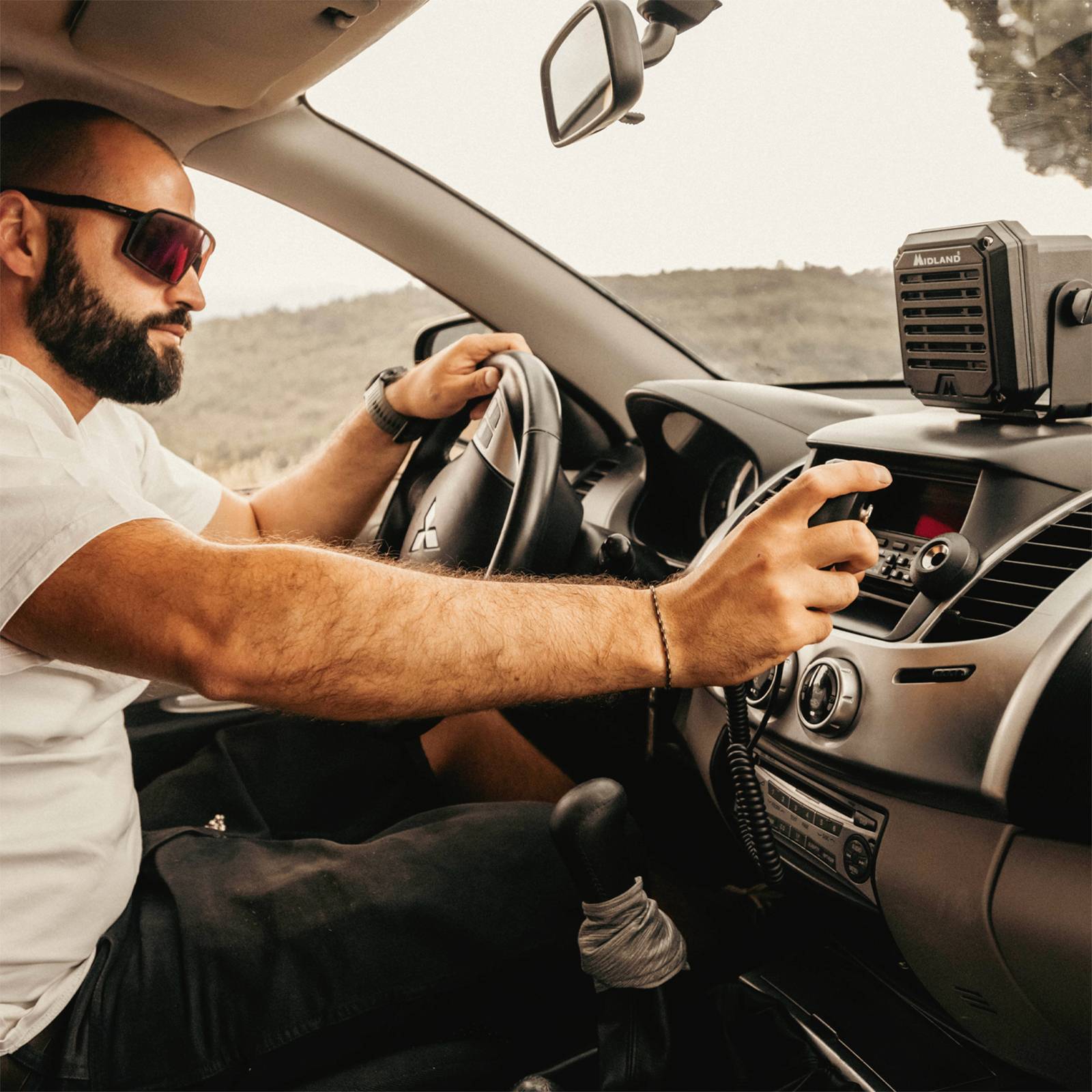 Ein Mann mit Sonnenbrille sitzt in einem Auto und bedient ein Funkgerät. Draußen ist eine Landschaft mit Hügeln zu sehen.