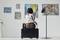 Eine Person sitzt auf einem Hocker in einer Kunstgalerie und betrachtet mehrere abstrakte Gemälde, die an einer weißen Wand hängen.