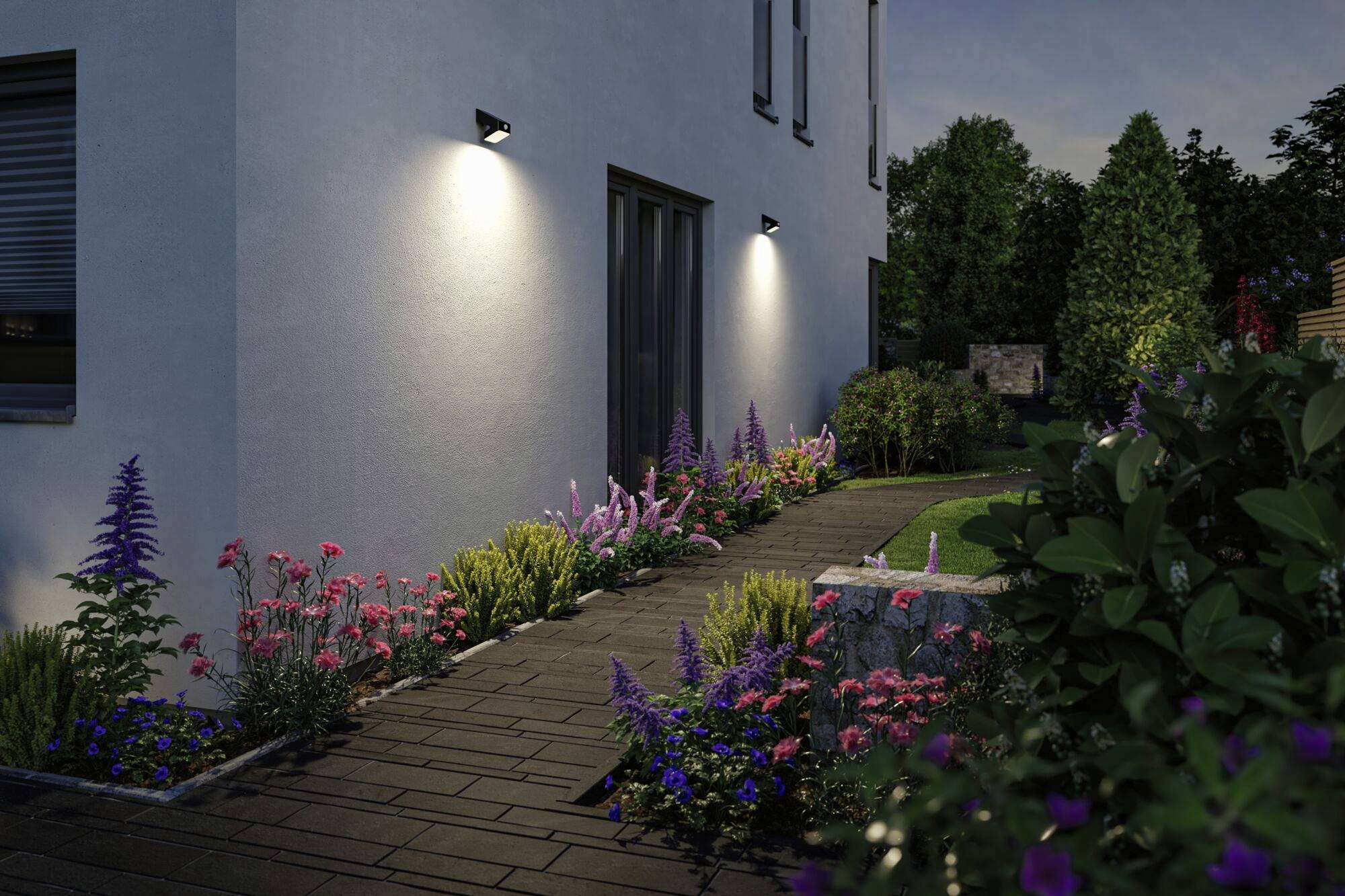 Ein beleuchteter Gartenweg mit bunten Blumenbeeten führt entlang eines modernen Hauses. Die Abendbeleuchtung erzeugt eine angenehme Atmosphäre.