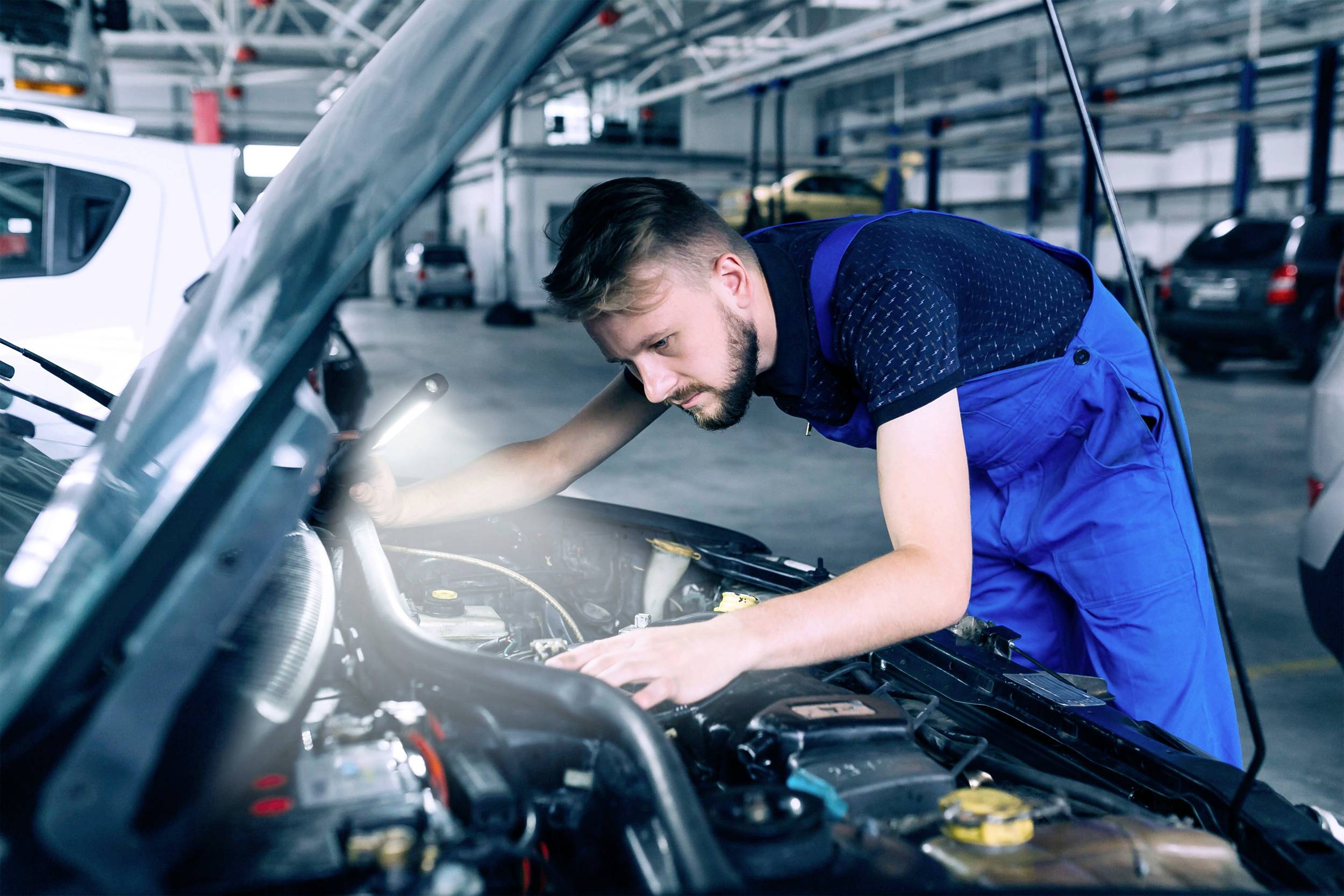 Ein Mechaniker in blauer Arbeitskleidung arbeitet konzentriert am Motor eines Autos in einer Werkstatt.