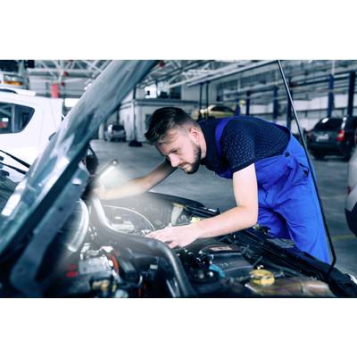 Ein Mechaniker in blauer Arbeitskleidung arbeitet konzentriert am Motor eines Autos in einer Werkstatt.