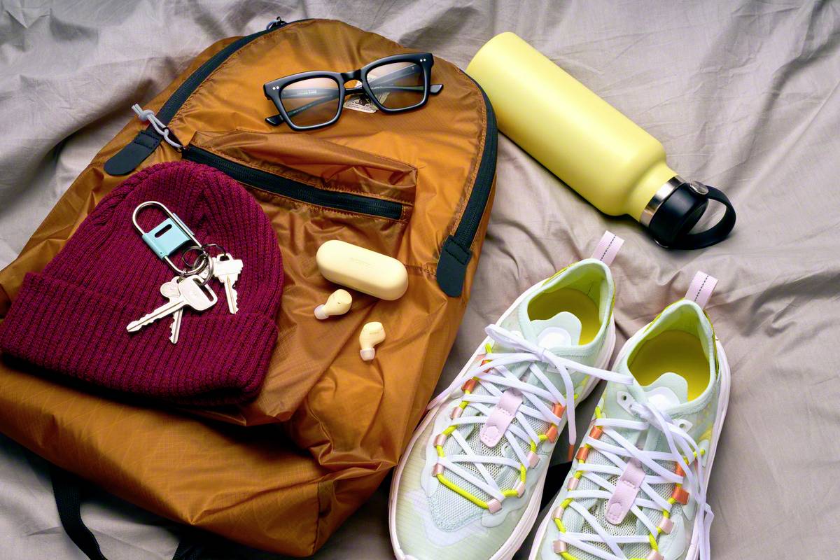 Ein orangefarbener Rucksack, Brille, gelbe Trinkflasche, rote Mütze mit Schlüsseln, kabellose Ohrstöpsel und helle Turnschuhe.