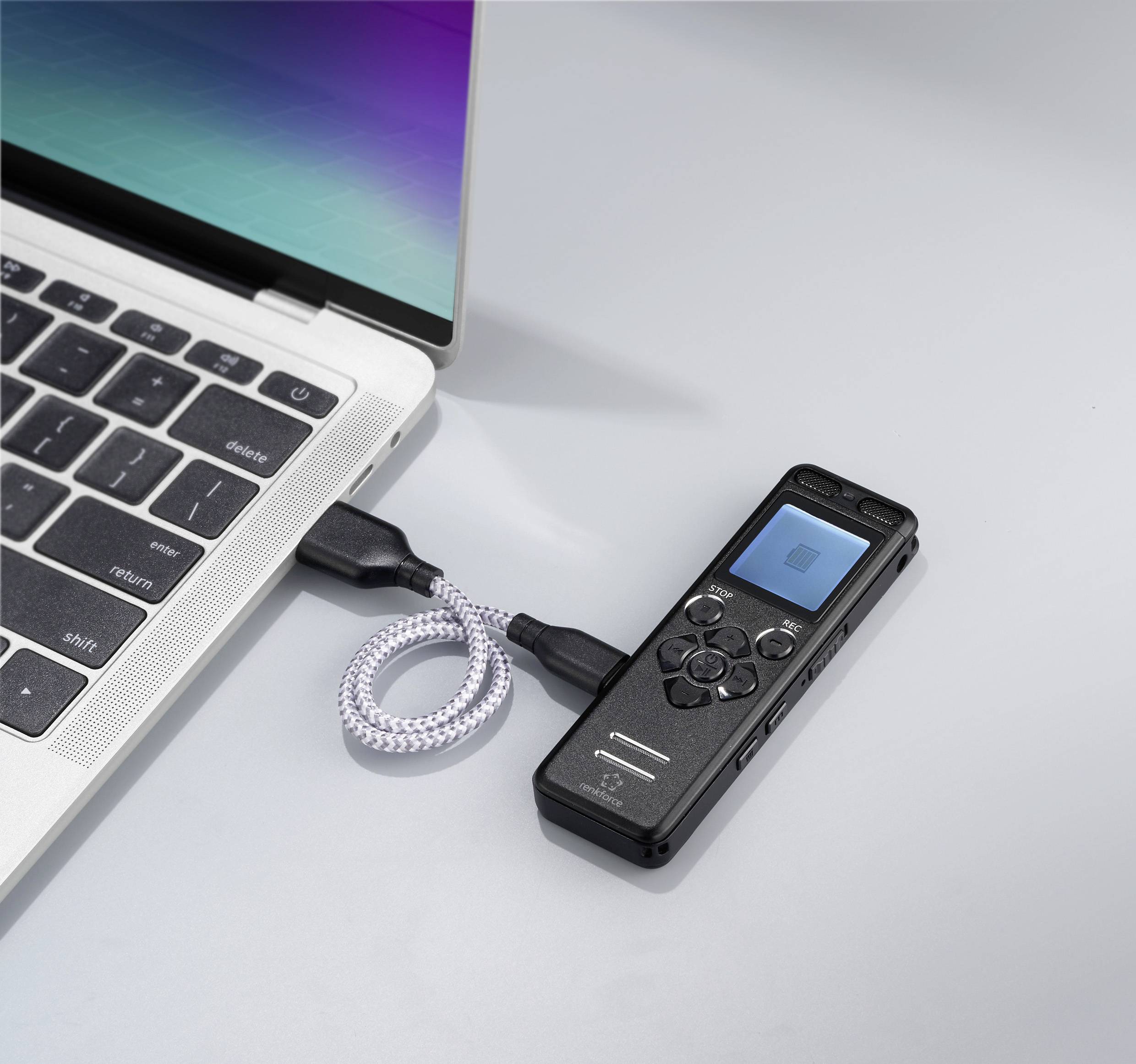 Ein Diktiergerät ist über ein Kabel mit einem Laptop verbunden. Der Laptop-Bildschirm zeigt ein Farbverlauf.