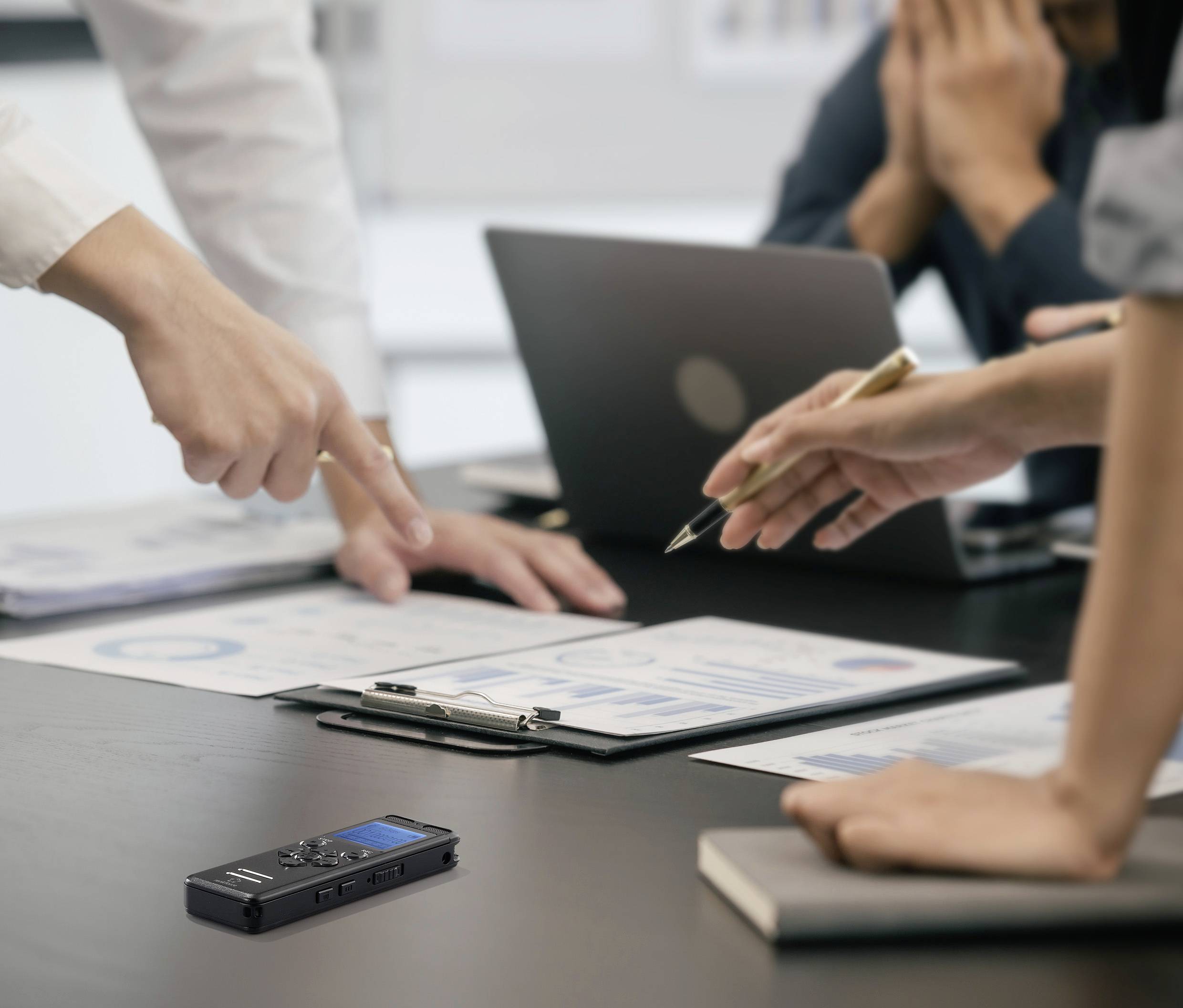Ein Geschäftstreffen, bei dem mehrere Personen über Dokumente auf einem Tisch diskutieren. Ein Diktiergerät liegt auf dem Tisch.