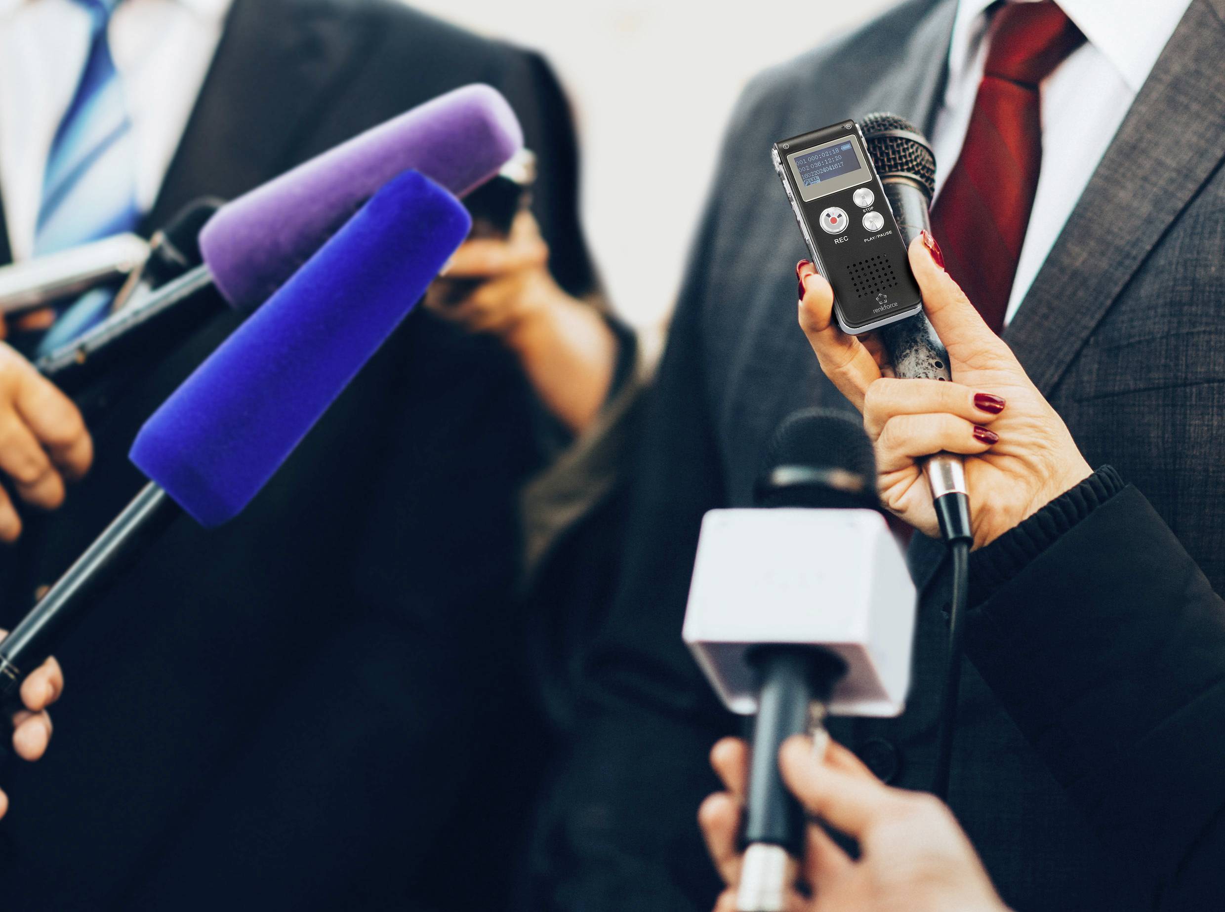 Person in Anzug spricht in mehrere Mikrofone bei einer Pressekonferenz. Hände halten blaue, violette und schwarze Mikrofone.