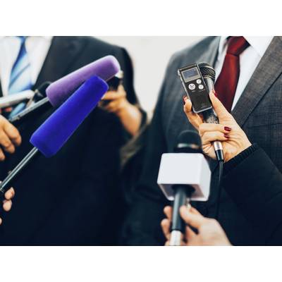 Person in Anzug spricht in mehrere Mikrofone bei einer Pressekonferenz. Hände halten blaue, violette und schwarze Mikrofone.