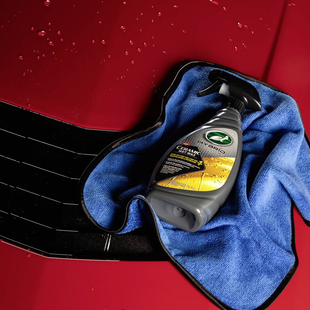 Flasche mit Keramik-Wachsspray auf blauem Tuch, platziert auf roter Motorhaube mit Wassertropfen. Pflegeprodukt für Autolack.