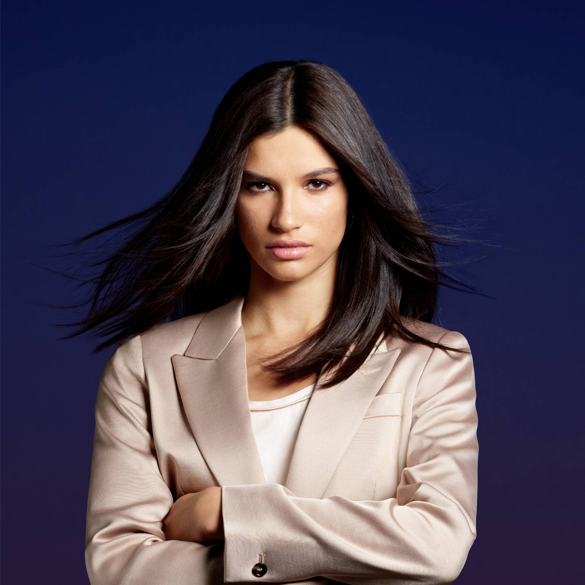 Eine Frau mit glattem, dunklem Haar trägt einen beigefarbenen Blazer vor einem dunkelblauen Hintergrund, schaut selbstbewusst.