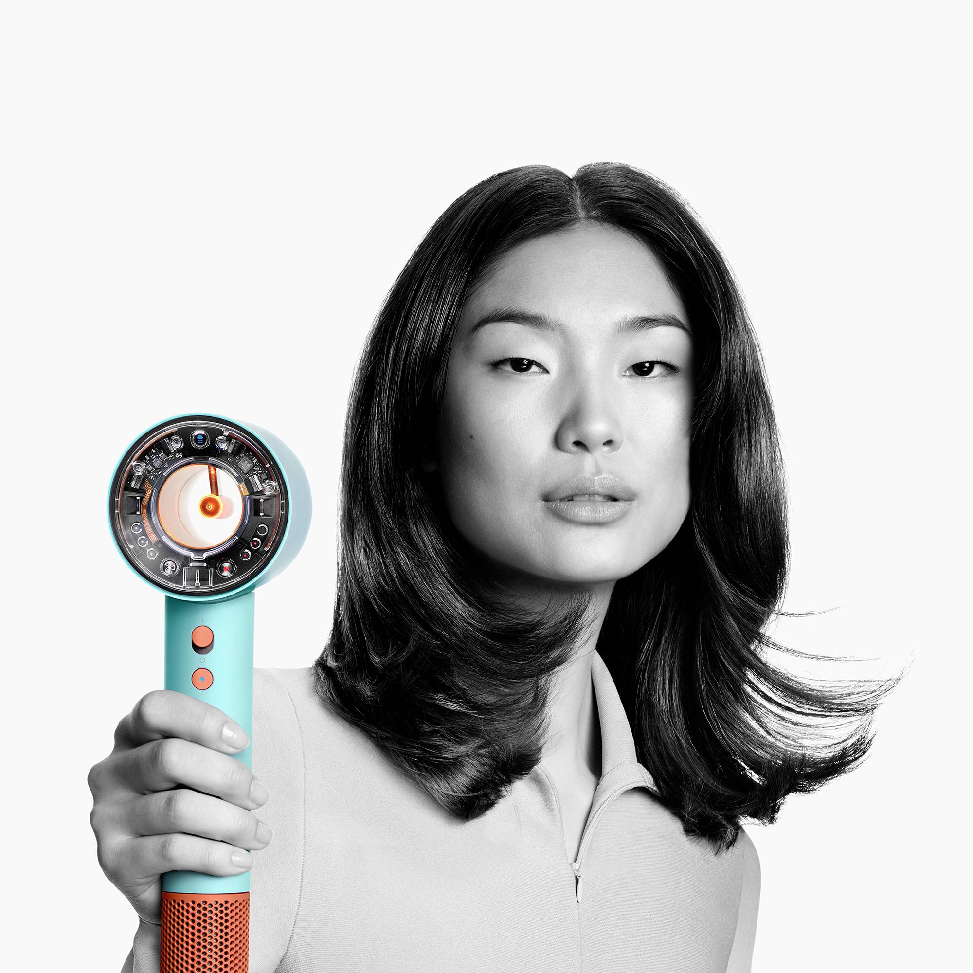 Zwei Personen mit lockigem Haar benutzen einen goltorangefarbenen Föhn mit türkisfarbenem Griff. Sie tragen identische graue Oberteile.