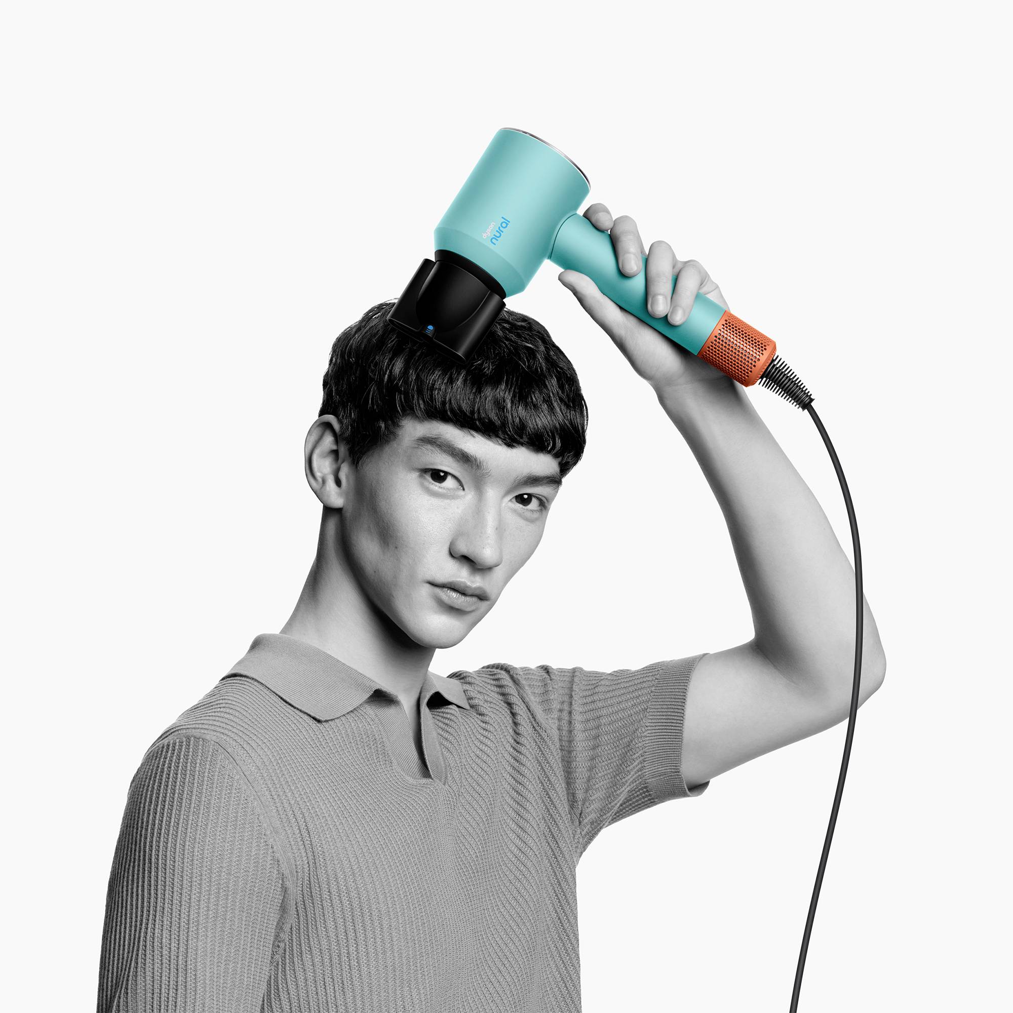 Eine Person mit einem elektrischen Haartrockner stylt das Haar, trägt ein helles Oberteil. Der Föhn ist blau und orange.