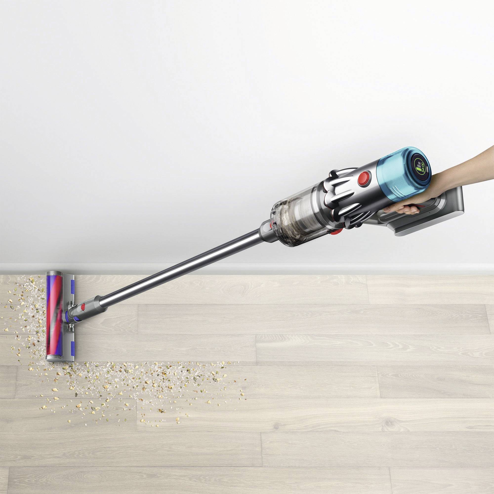 Ein Staubsauger mit einem durchsichtigen Staubbehälter reinigt Müsli, das auf einem Holzboden verstreut ist. Eine Hand einer Person umgreift den Griff.