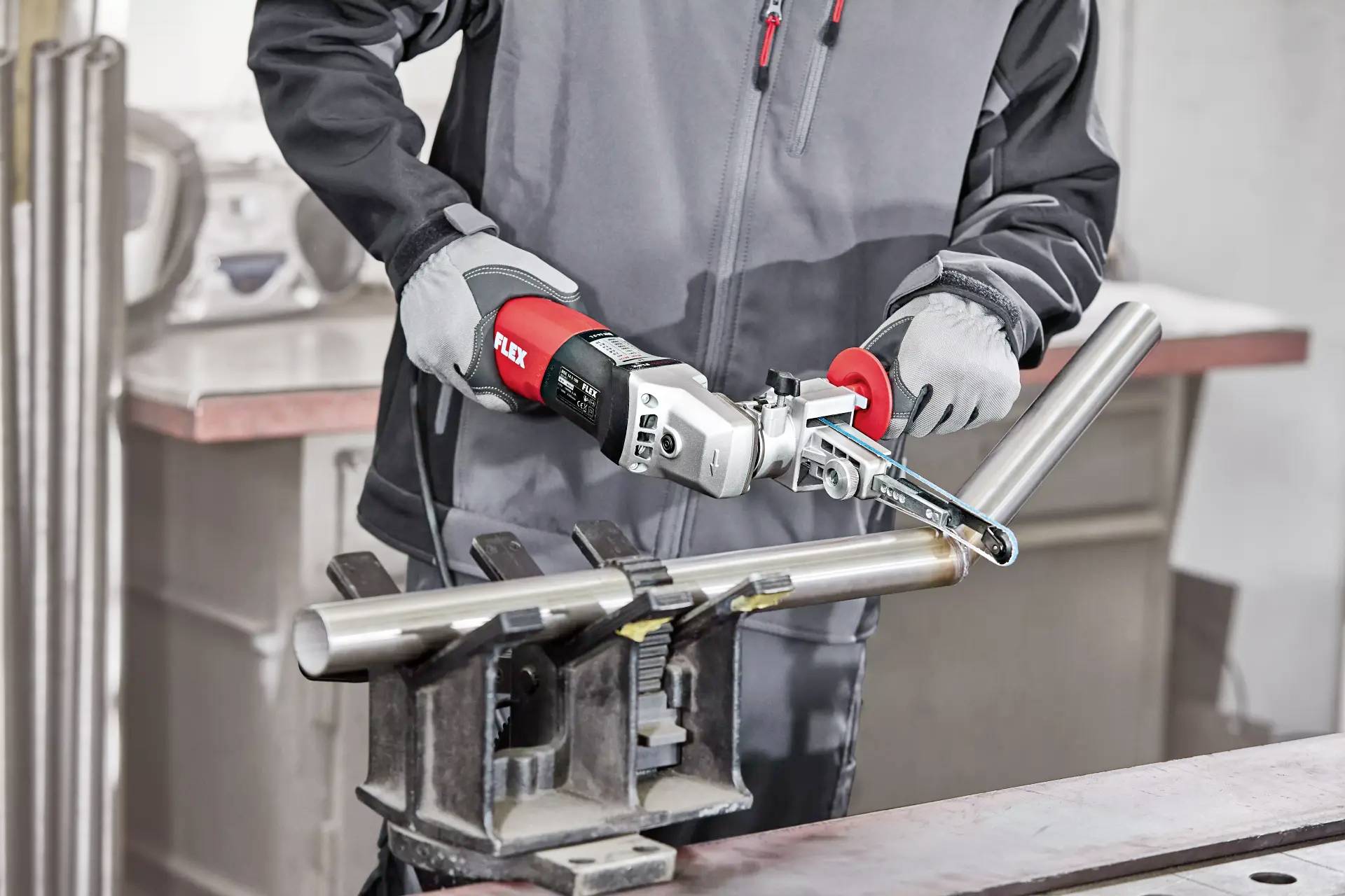 Eine Person arbeitet mit einem Bandschleifer an einem Metallrohr in einer Werkstatt. Der Fokus liegt auf dem Werkzeug und der Metallbearbeitung.
