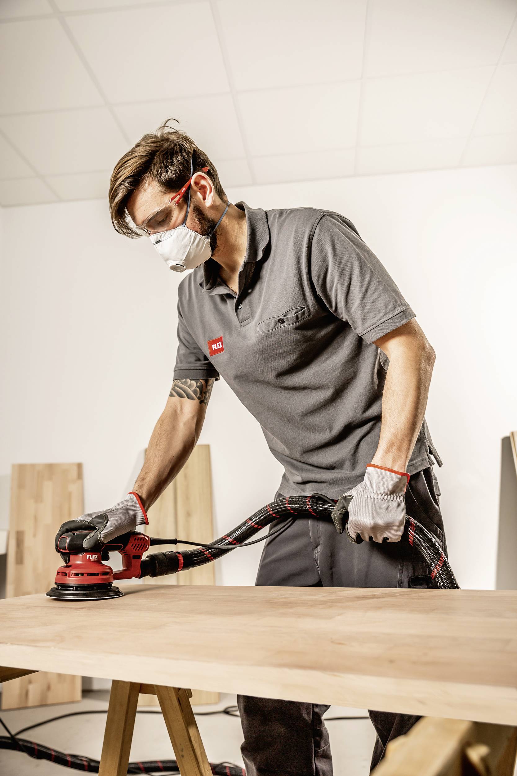 Ein Mann schleift mit einer Schleifmaschine eine Holzplatte. Er trägt Schutzbrille, Atemmaske und Handschuhe in einer Werkstattumgebung.