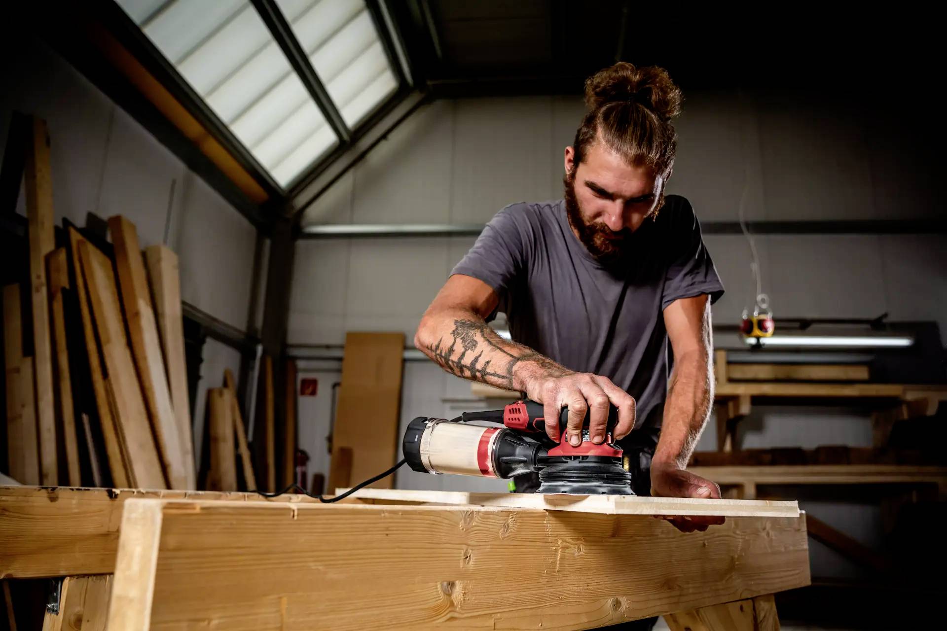 Ein Mann schleift konzentriert ein Holzbrett in einer Werkstatt mit einer elektrischen Schleifmaschine. Sonnenlicht dringt durch ein Fenster.