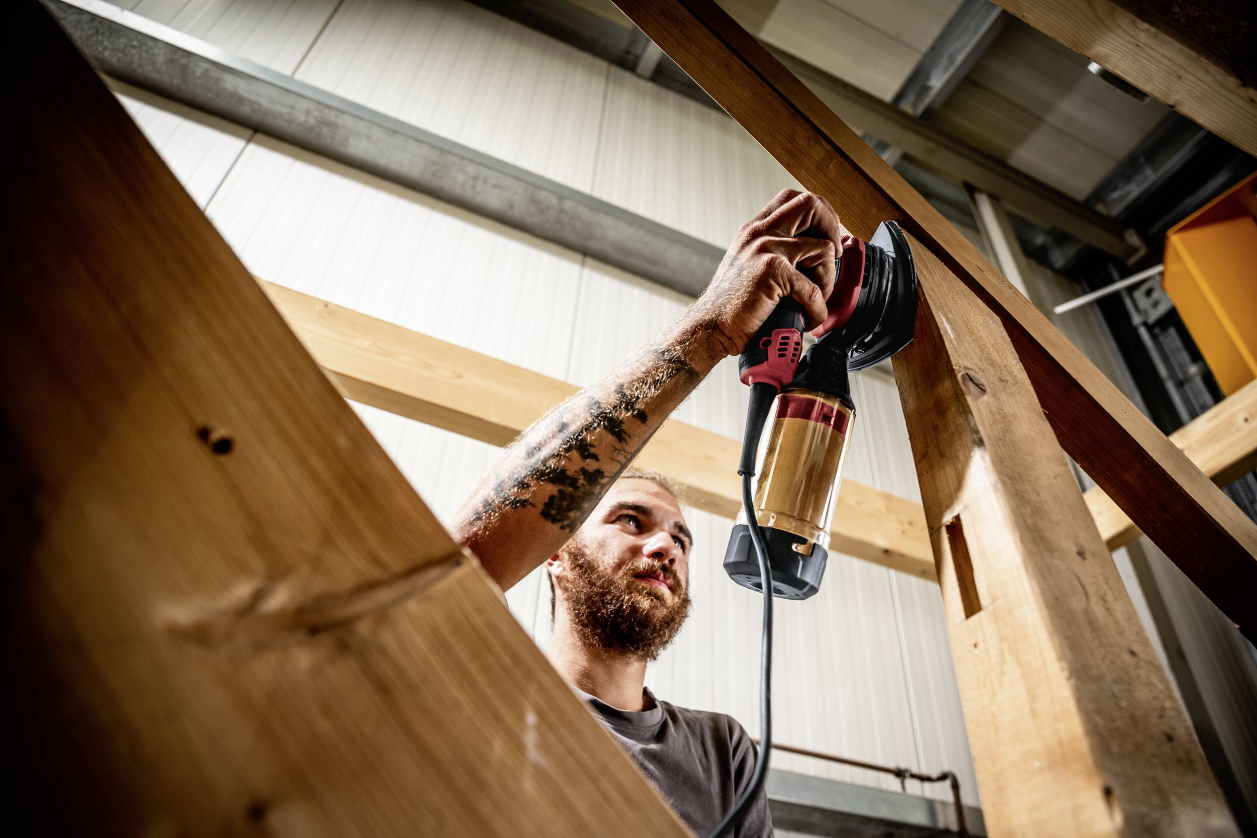 Ein Mann arbeitet in einer Werkstatt, verwendet eine elektrische Schleifmaschine an einem Holzbalken. Er konzentriert sich auf seine Aufgabe.