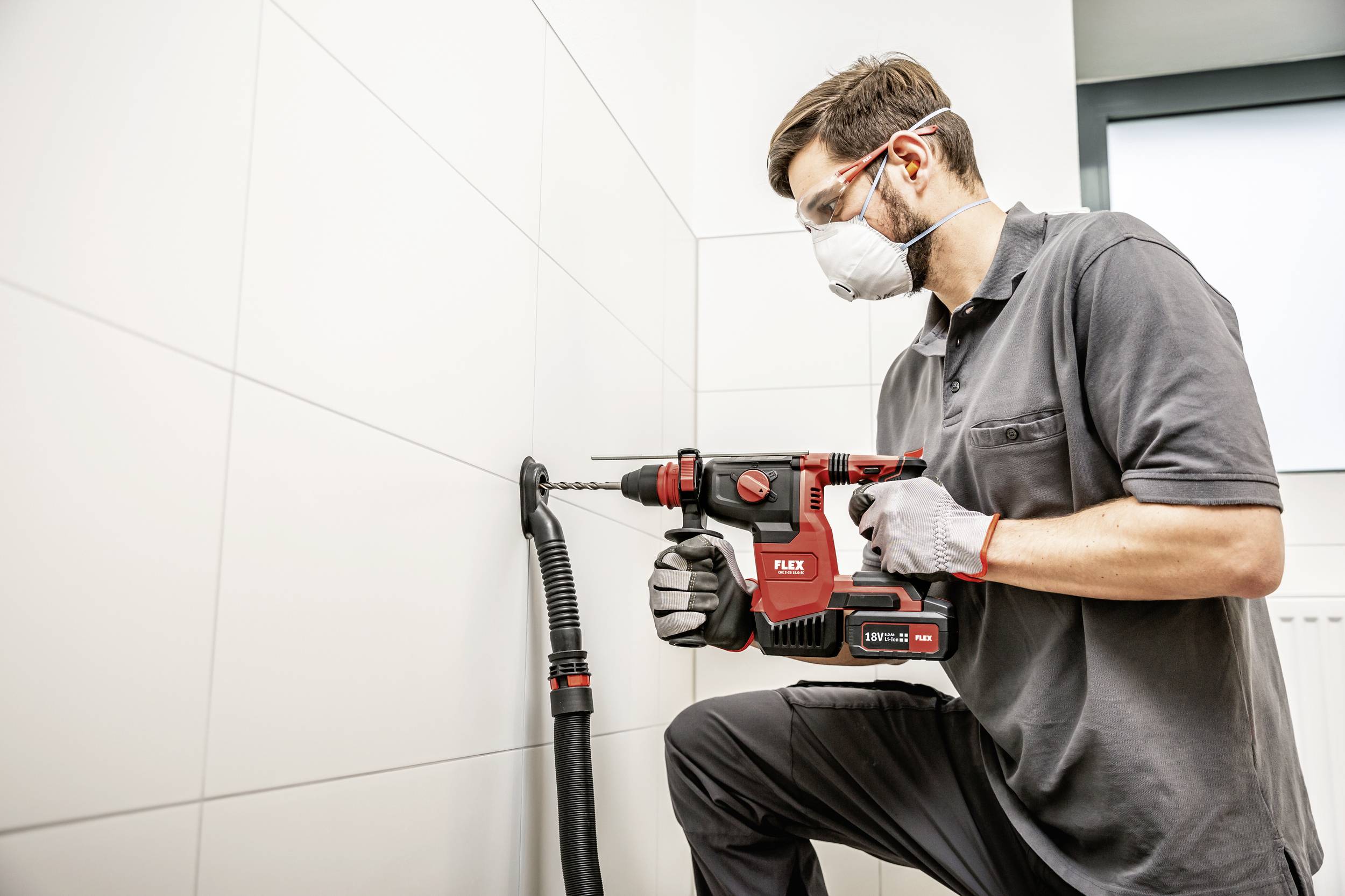 Ein Handwerker in Schutzmaske und Brille bohrt mit einem roten Bohrhammer ein Loch in eine geflieste Wand. Ein Staubsauger saugt den Bohrstaub ab.