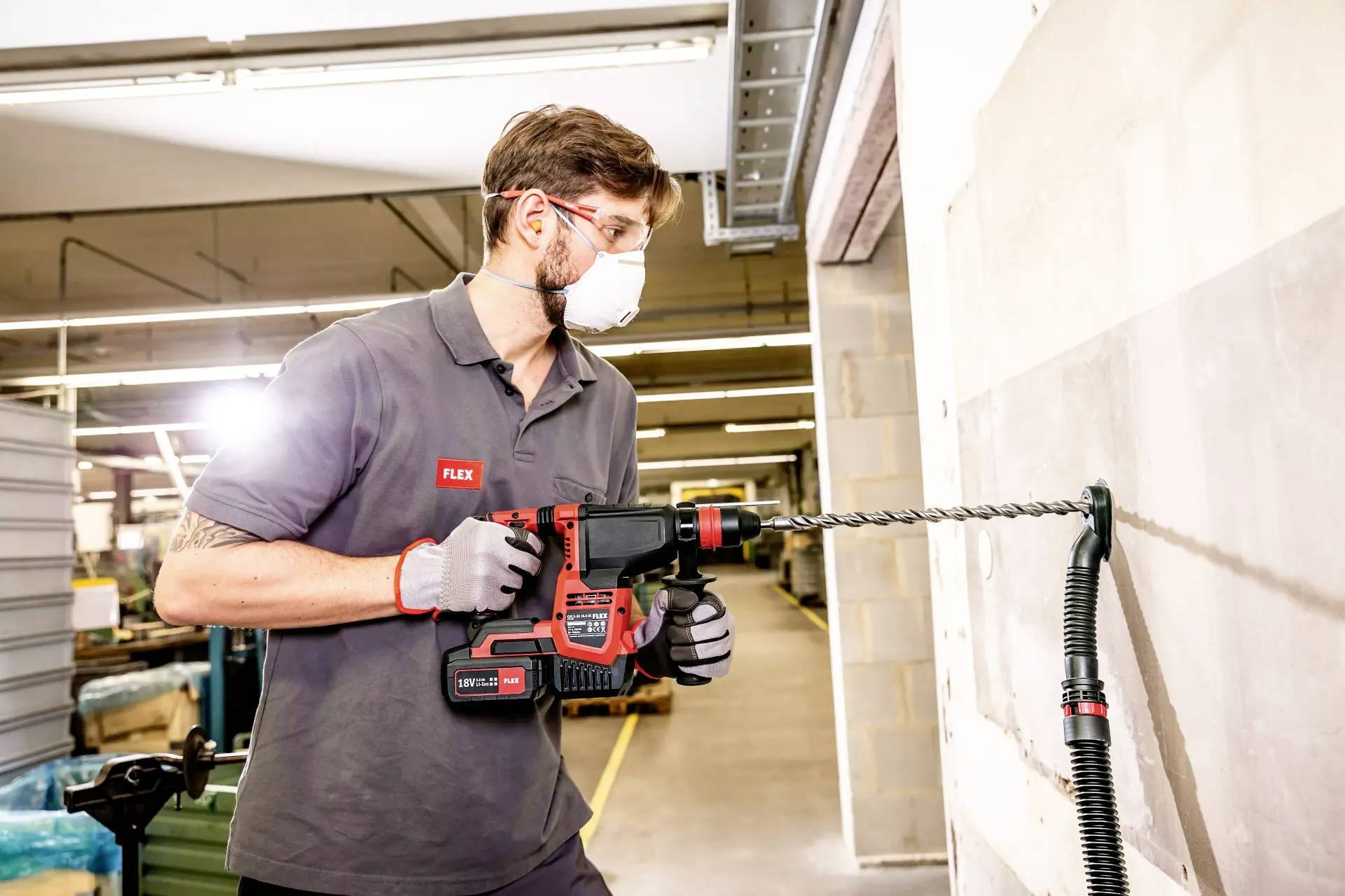 Ein Mann mit Schutzbrille und Maske bohrt mit einem schweren Bohrhammer in eine Wand in einer Werkhalle, umgeben von Ausrüstung.